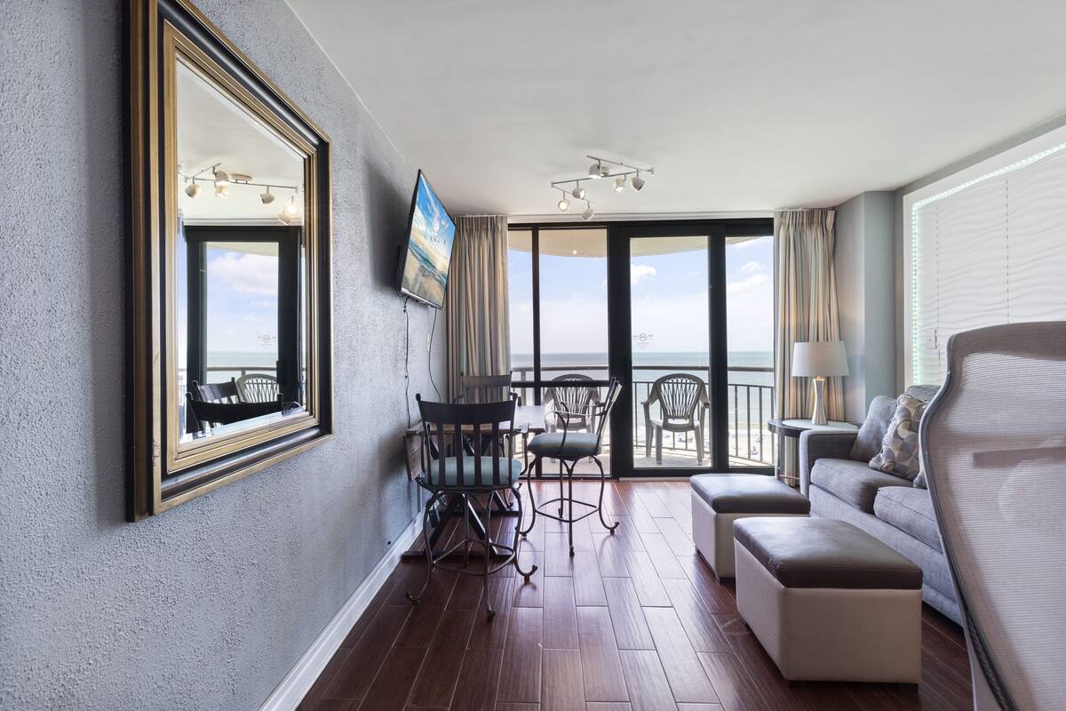 Habitación de hotel con balcón, sofá, mesa de comedor con sillas y un espejo grande.