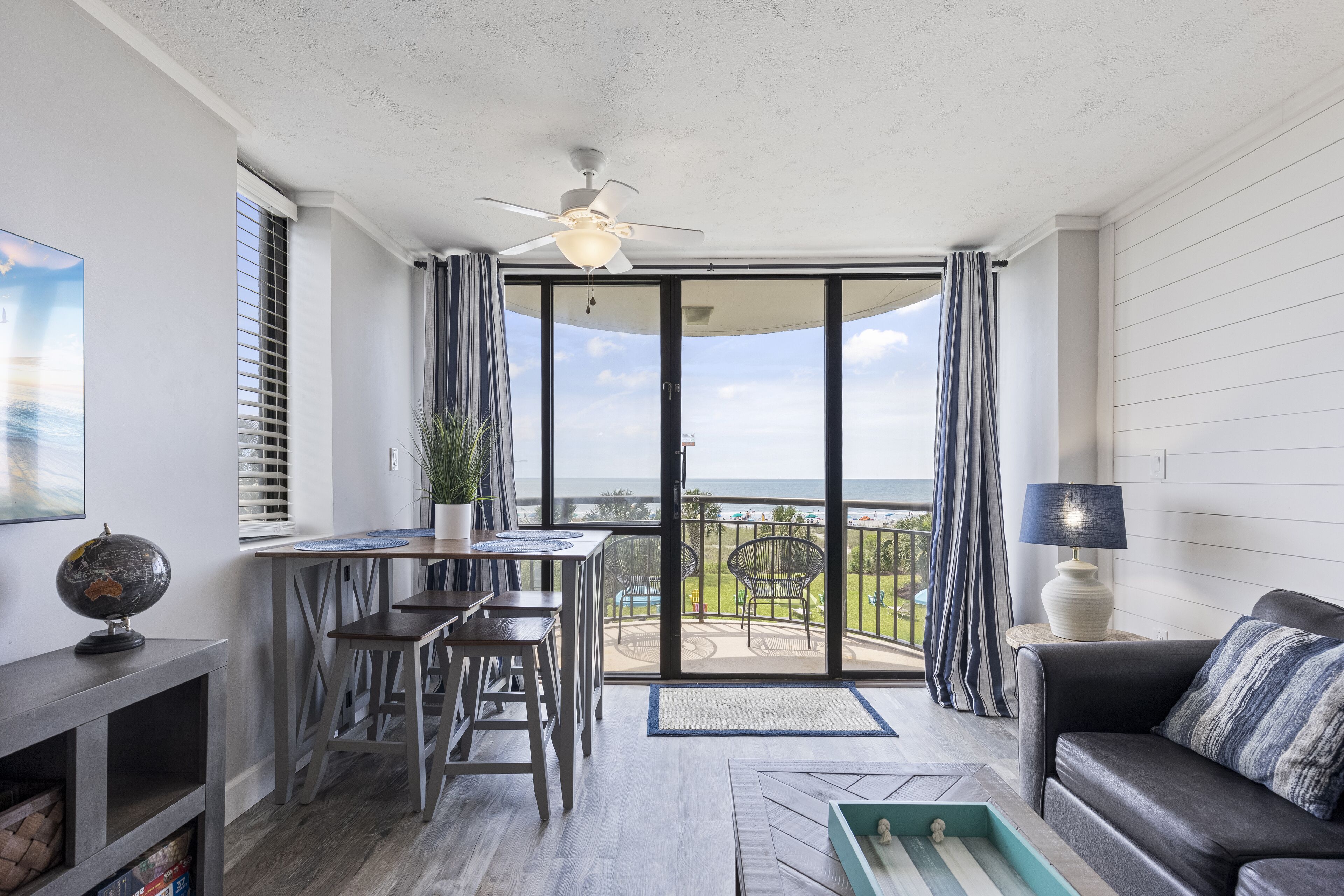 A modern living room with a balcony, a ceiling fan, a dining area with stools, and a sofa with a striped cushion.