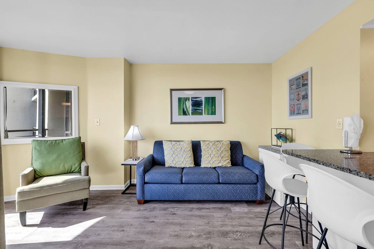 Una sala de estar con un sofá azul, un sillón verde y una zona de bar blanca con taburetes.