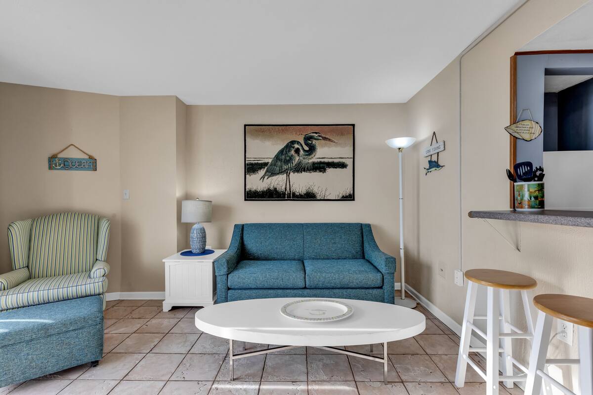 Una sala de estar con un sillón a rayas, un sofá azul, una mesa de centro blanca y un cuadro de un pájaro.