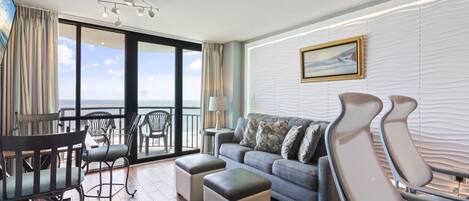 Una sala de estar moderna con un sofá, otomanas y sillones, un balcón con vistas, y un cuadro en la pared.