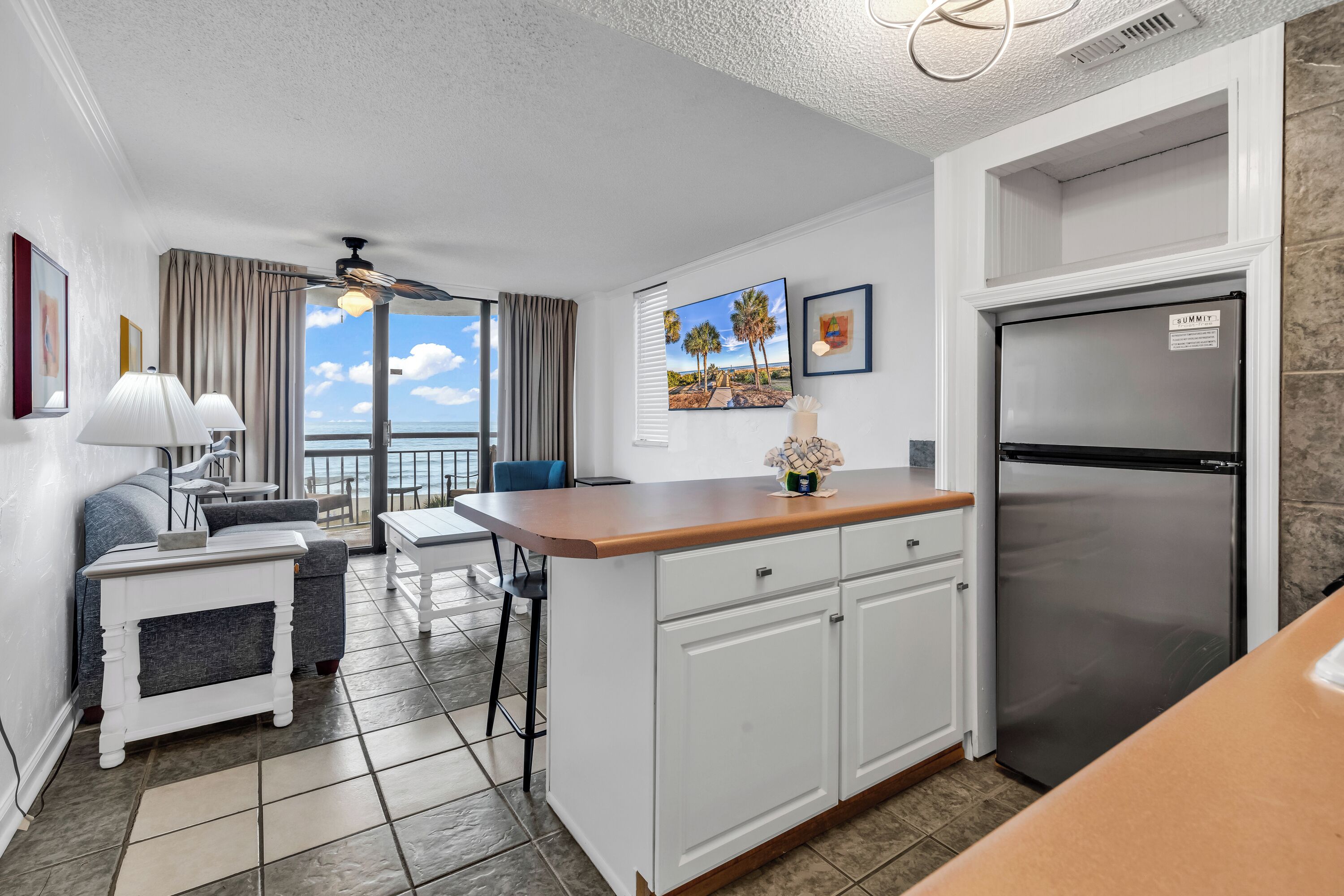 A compact kitchen with a refrigerator, a dining area with a table and chairs, and a view of the ocean through large windows.