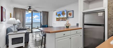 A compact kitchen with a refrigerator, a dining area with a table and chairs, and a view of the ocean through large windows.