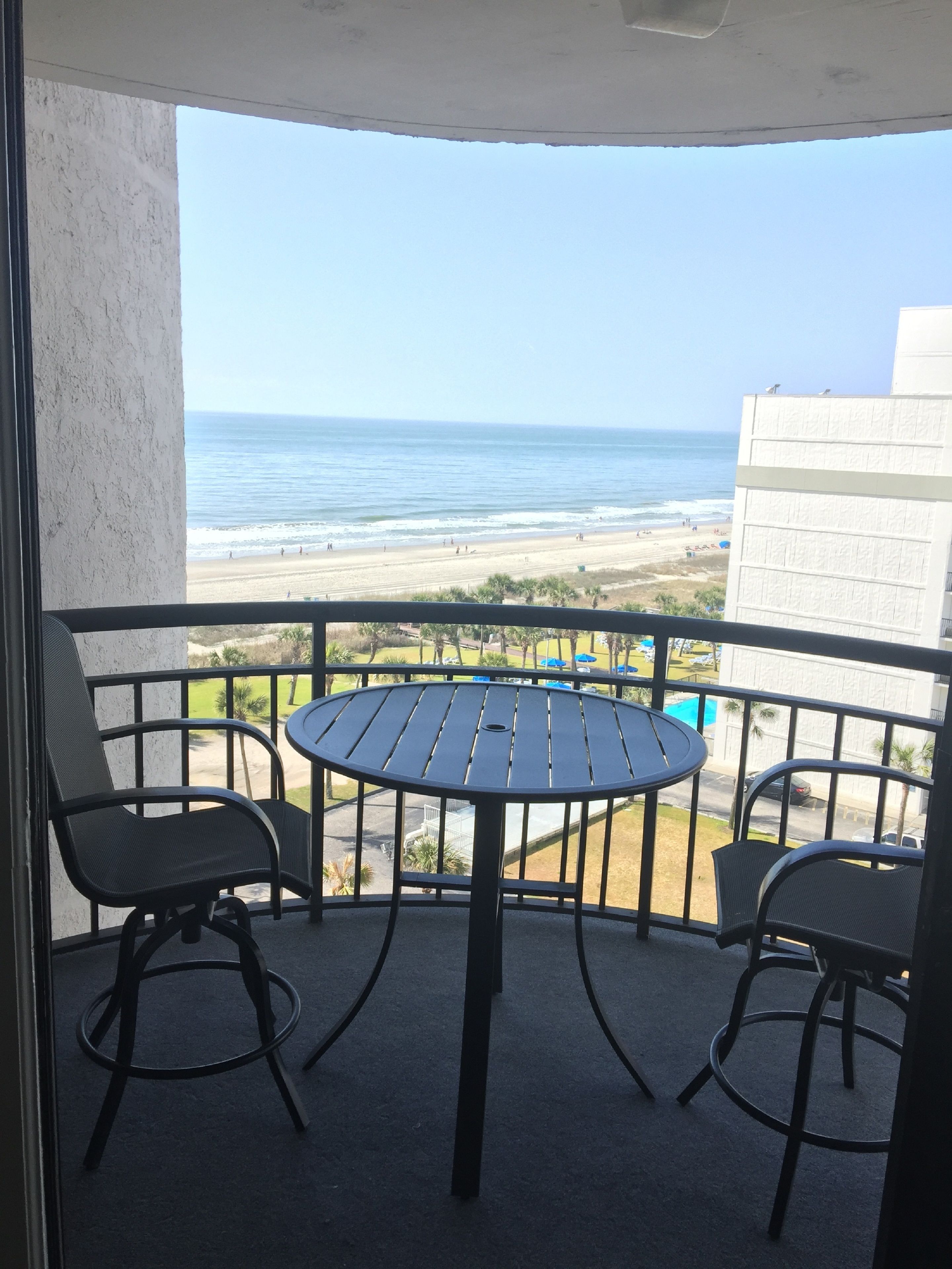 A balcony with a round table and two chairs, overlooking a beach with people and a clear sky.