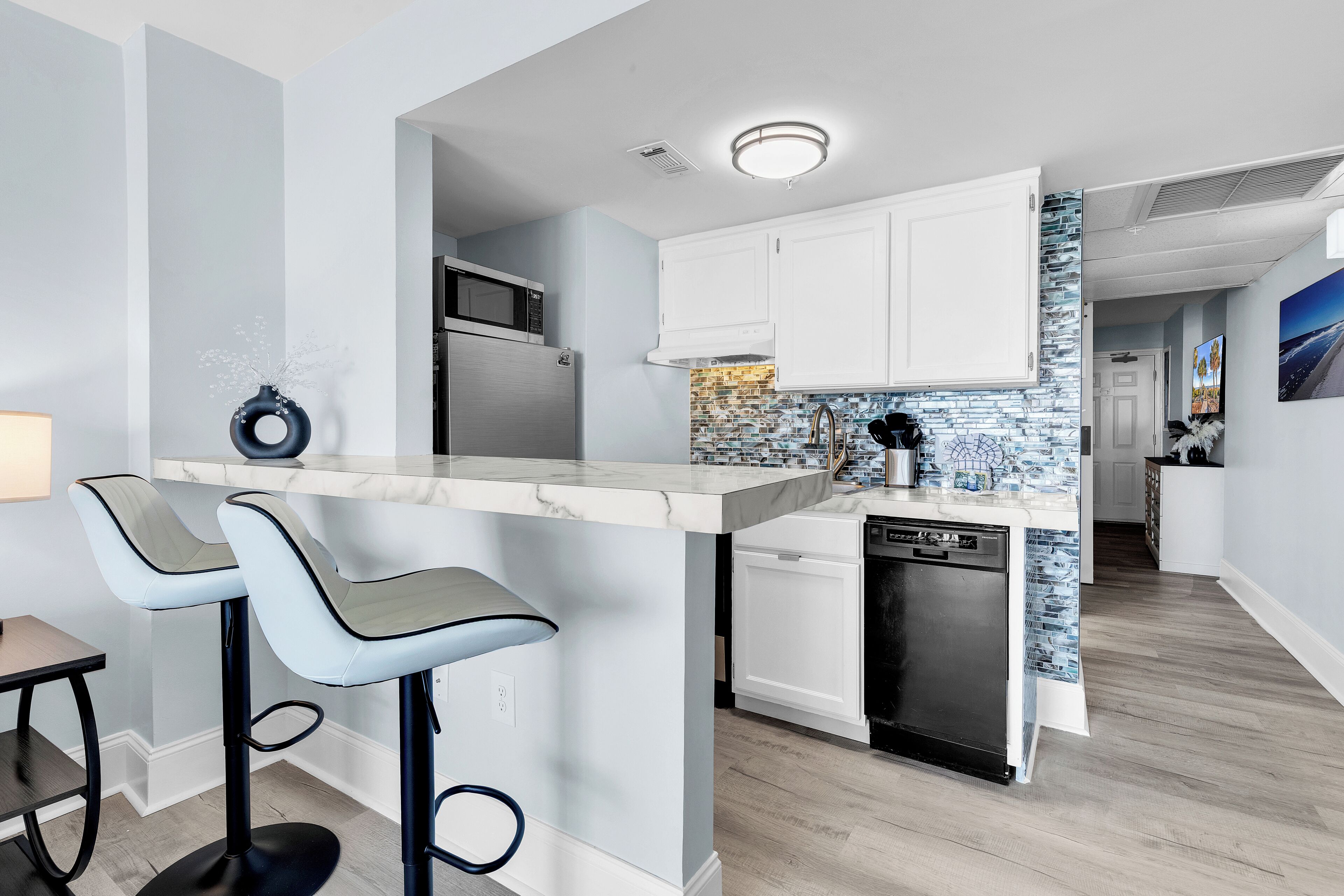 A modern kitchen with a marble island, bar stools, a dishwasher, and a tiled backsplash.