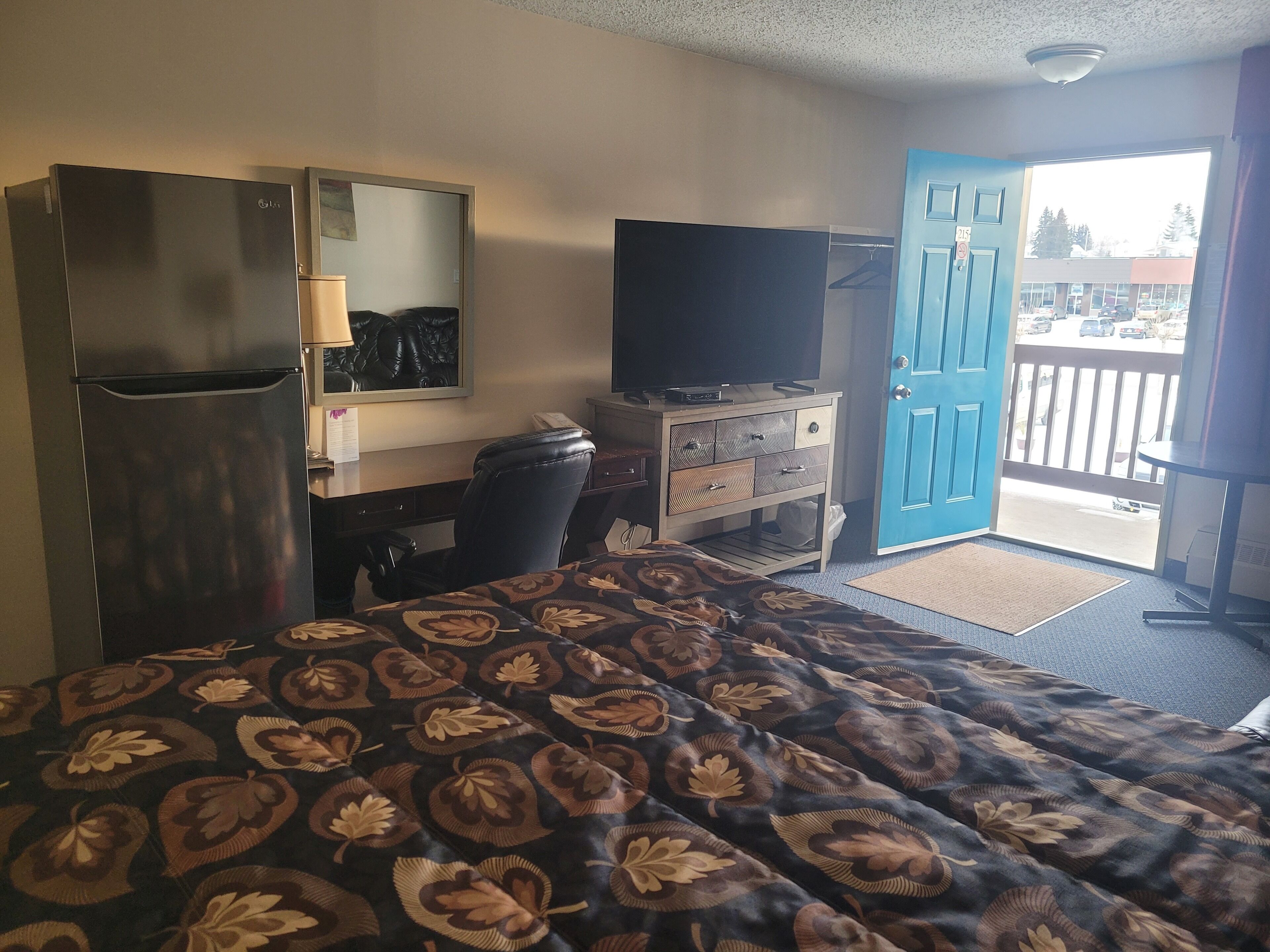 A hotel room with a bed, a desk, a television, a refrigerator, and a blue door leading to a balcony.