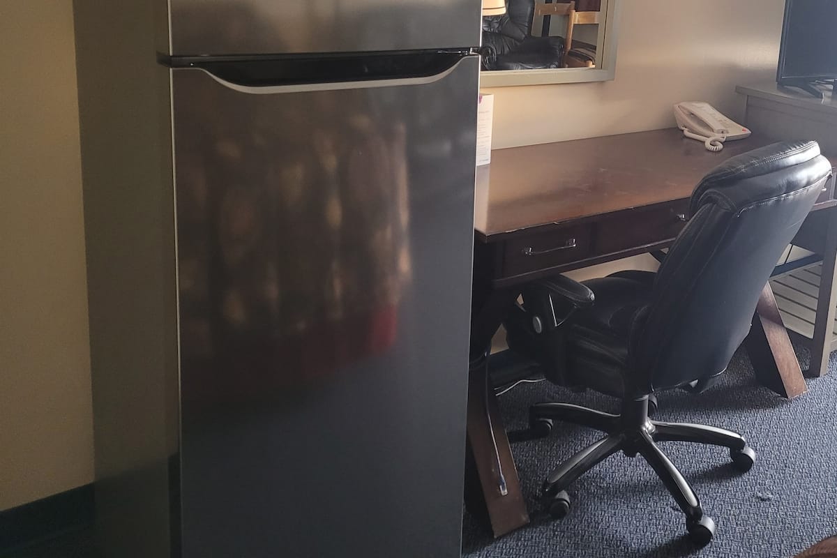 A hotel room with a refrigerator, a desk, a chair, a lamp, and a telephone.