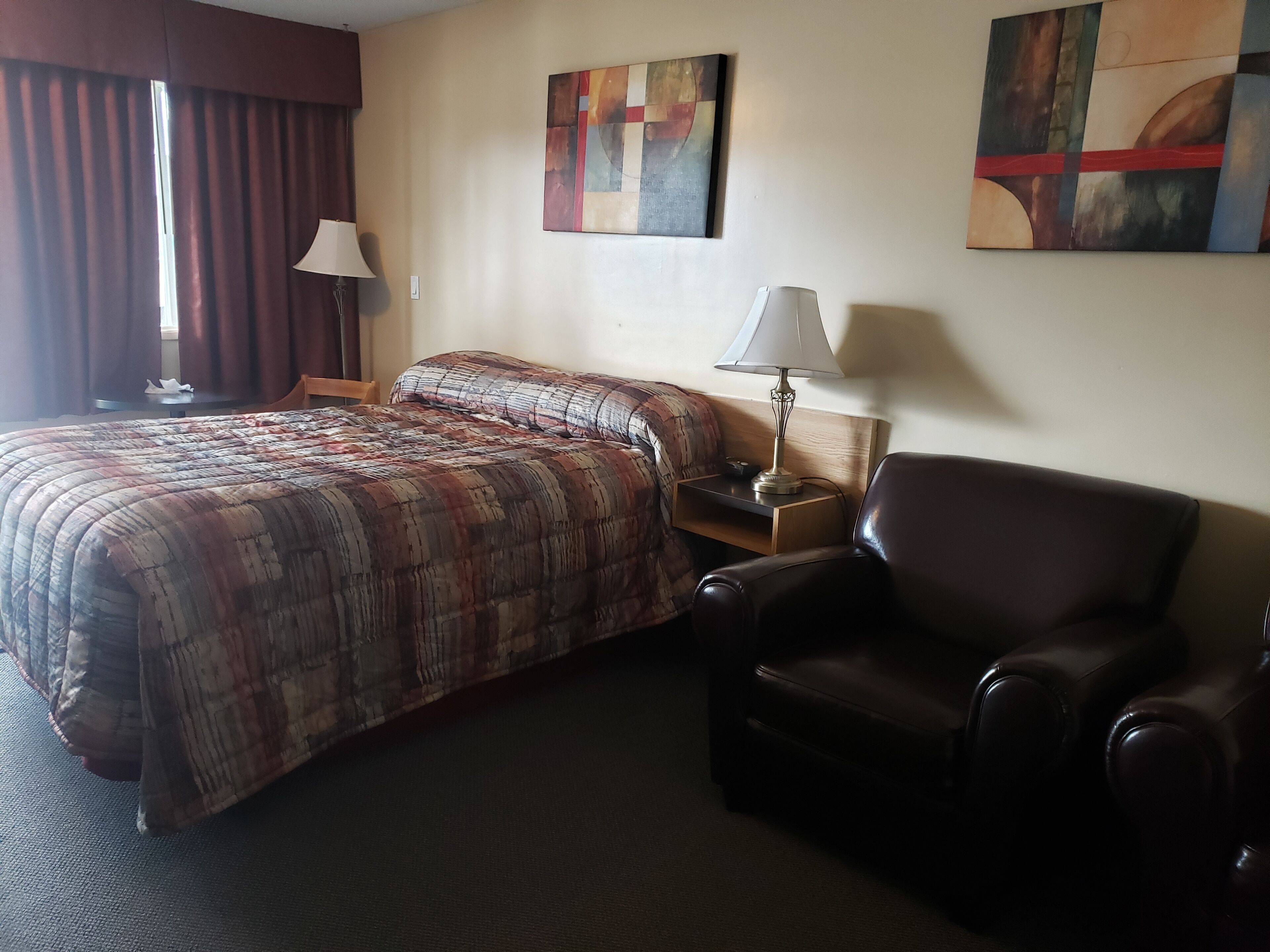 Une chambre d’hôtel avec un lit, une chaise, une table de chevet, une lampe et deux tableaux abstraits accrochés au mur.