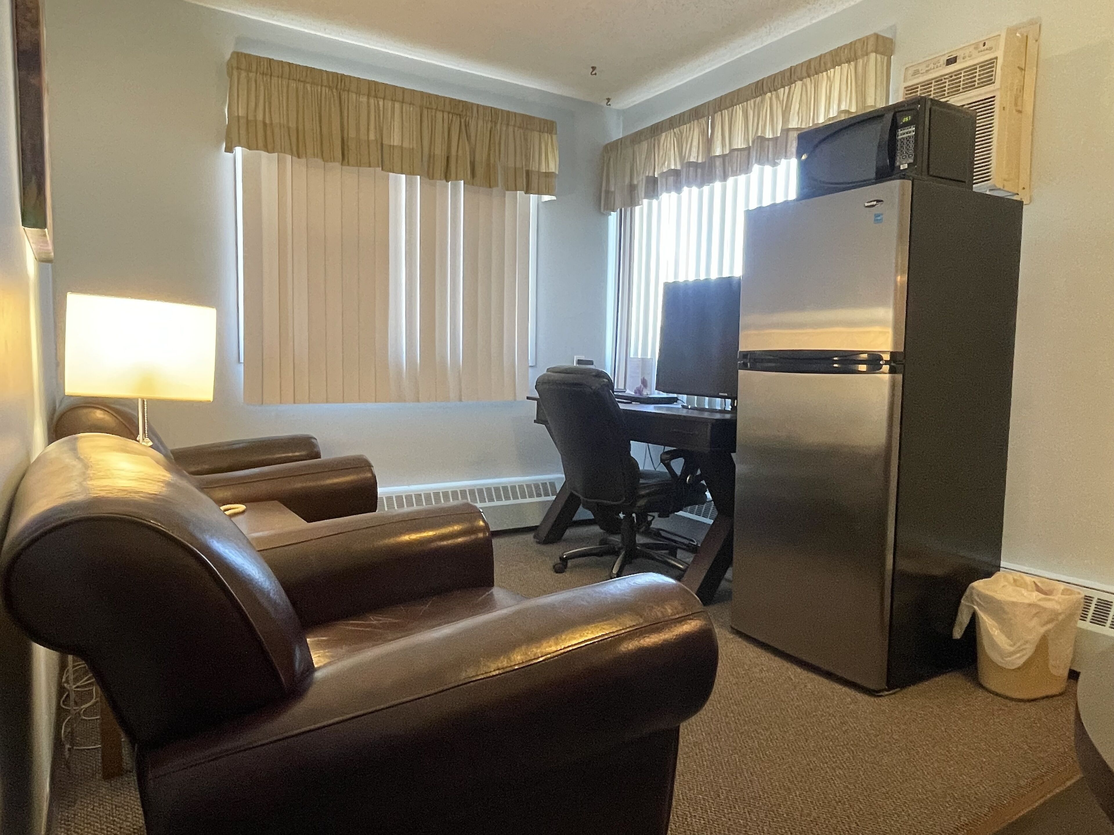 A hotel room with a leather sofa, a desk with a computer, a refrigerator, and a window with curtains.