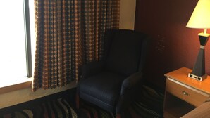 Habitación de hotel con una sola silla, una mesita de noche con lámpara y una ventana con cortinas estampadas.