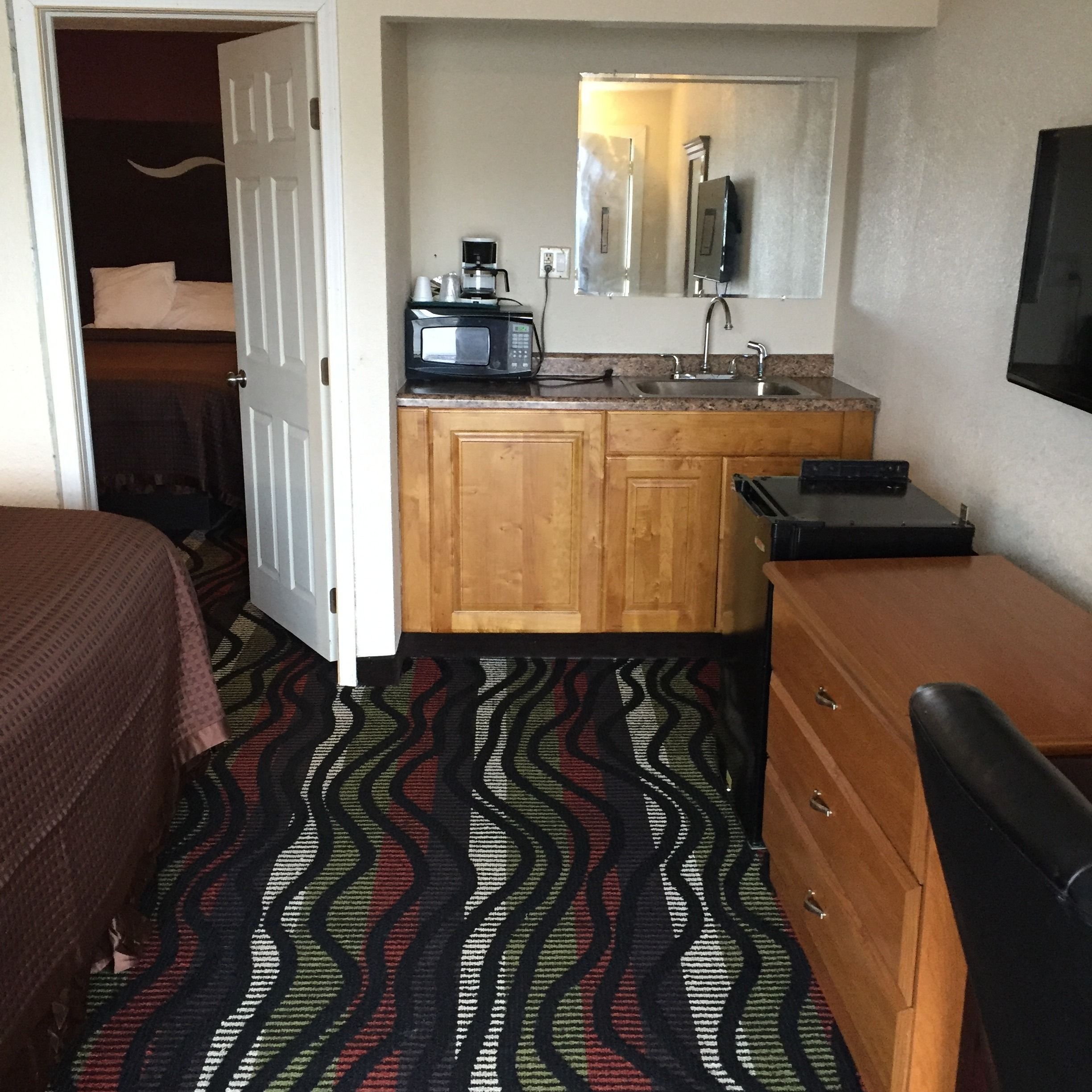 Une chambre d’hôtel compacte comprenant un coin cuisine, un lit avec une couvre-lit à motifs, une commode en bois et un téléviseur fixé au mur.