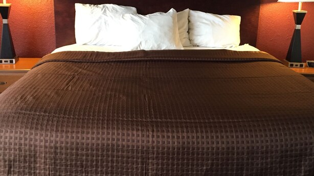 Una cama bien hecha con una manta marrón y almohadas blancas, flanqueada por dos mesitas de noche negras con lámparas.