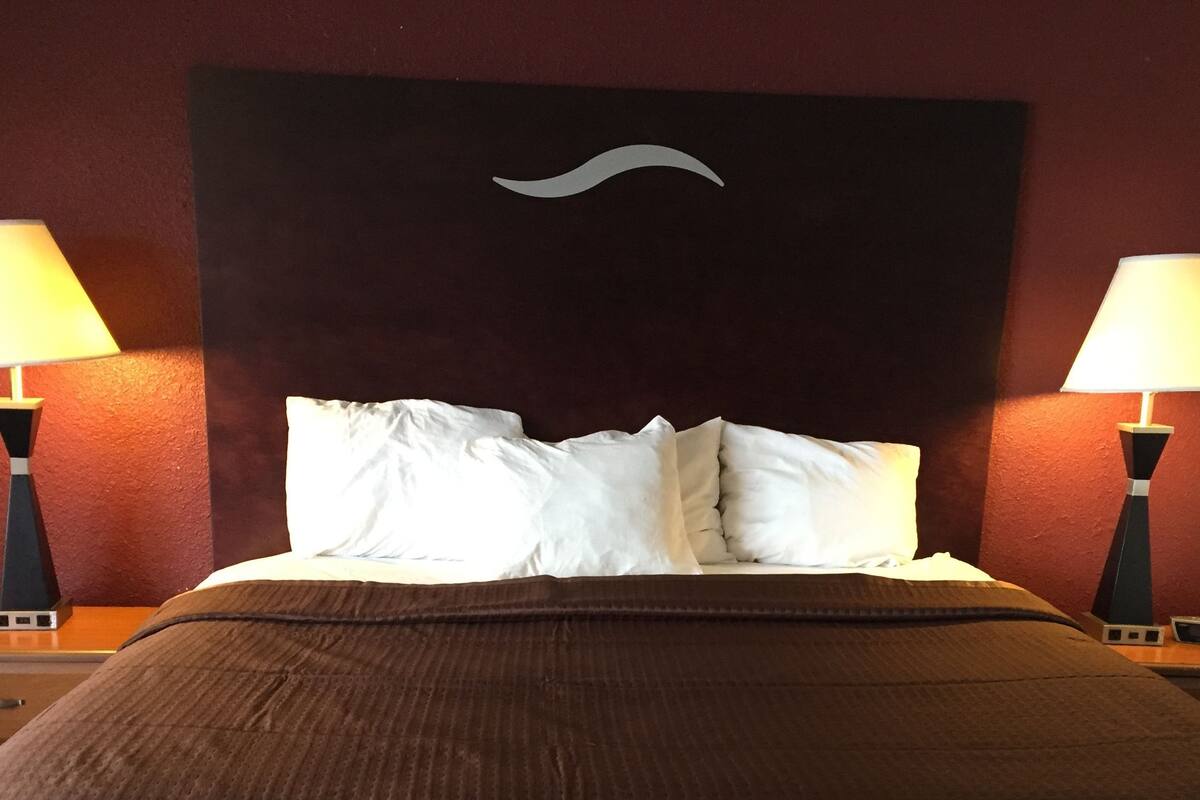 Un lit bien fait, recouvert d’une couverture marron et agrémenté d’oreillers blancs, avec deux tables de chevet noires de chaque côté, sur lesquelles sont posées des lampes.