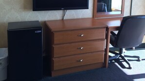 Habitación de hotel con televisor de pantalla plana montado en la pared, cómoda de madera con cajones, refrigerador negro pequeño, silla de oficina negra y un espejo sobre la cómoda.