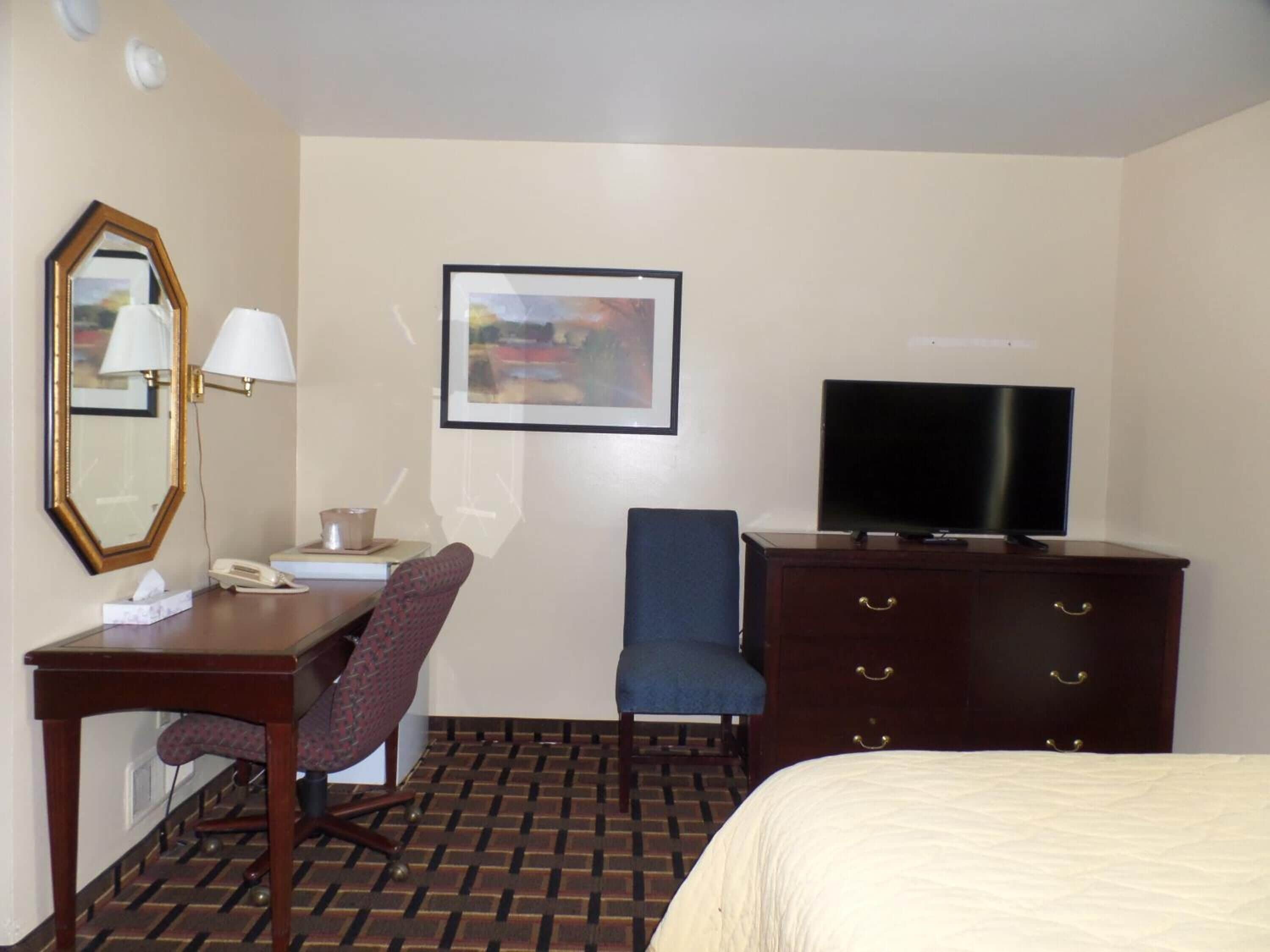 Ein Hotelzimmer mit einem Bett, einem Schreibtisch mit Telefon, einem Sessel, einer Kommode, einem Fernseher, einem gerahmten Bild an der Wand und einem gemusterten Boden.
