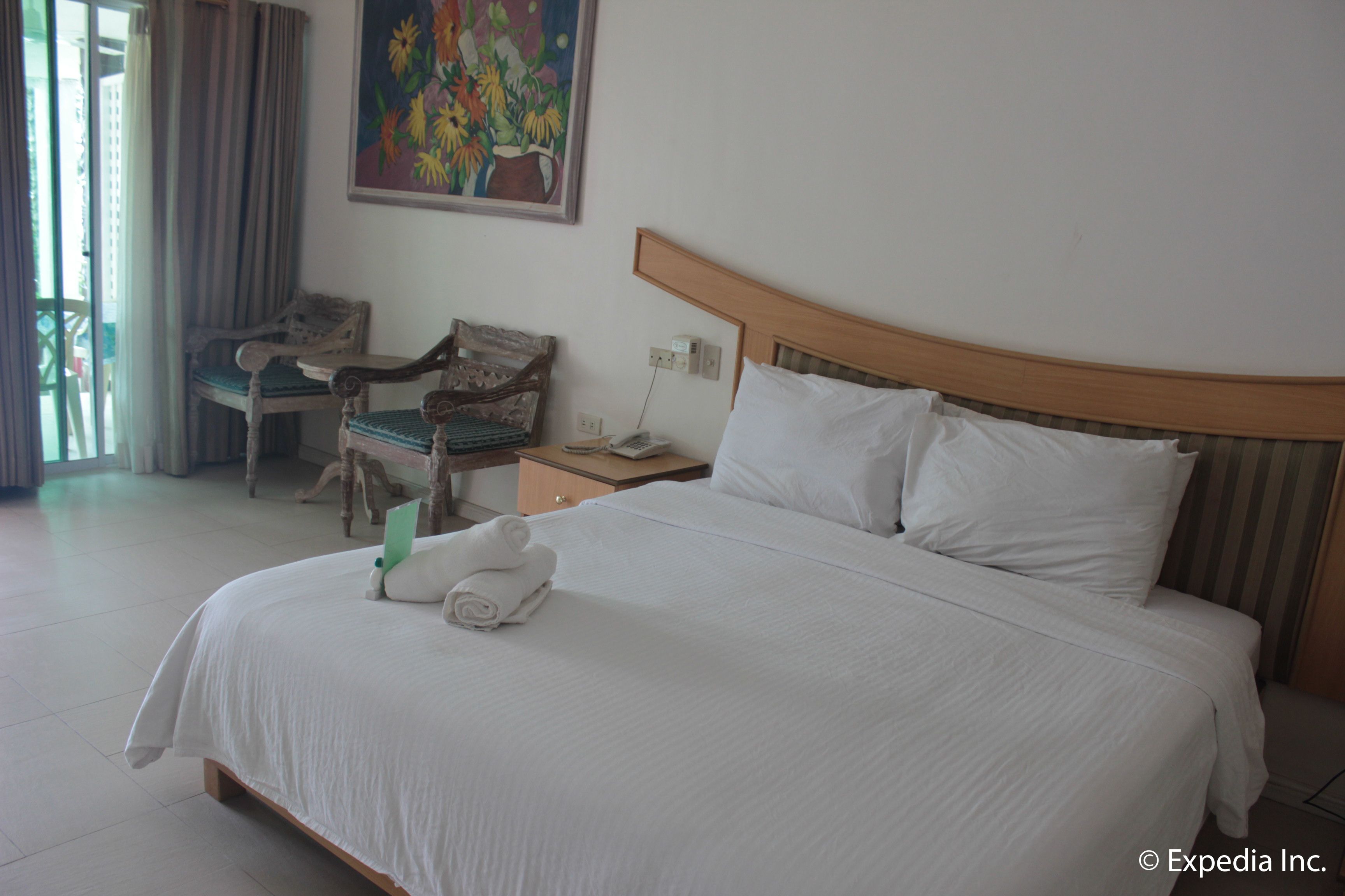 A neatly made bed with white linens and pillows, a wooden headboard, and a painting of flowers on the wall.