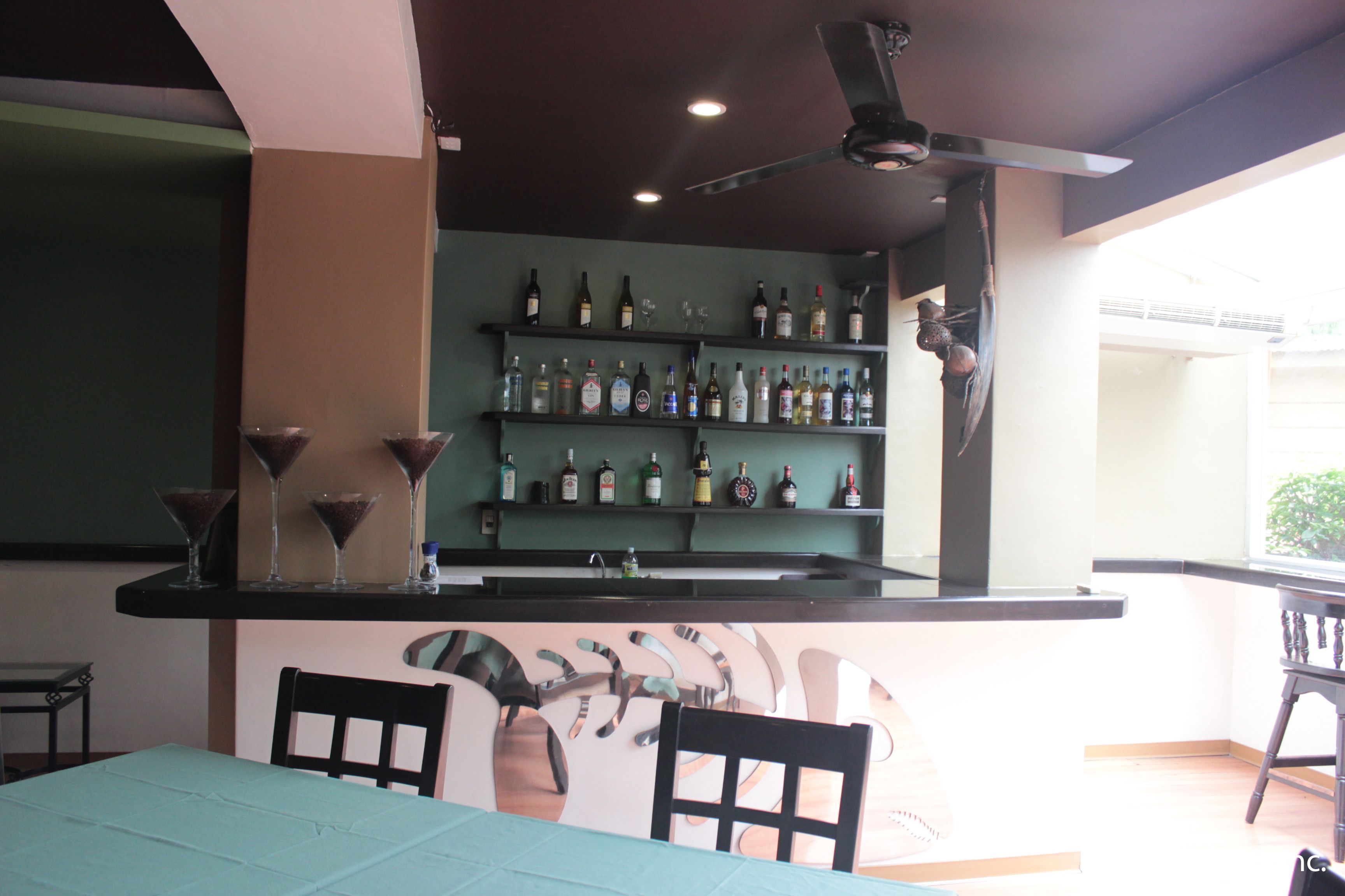 Zona de bar con estantes llenos de botellas de licores, una barra y un ventilador de techo.