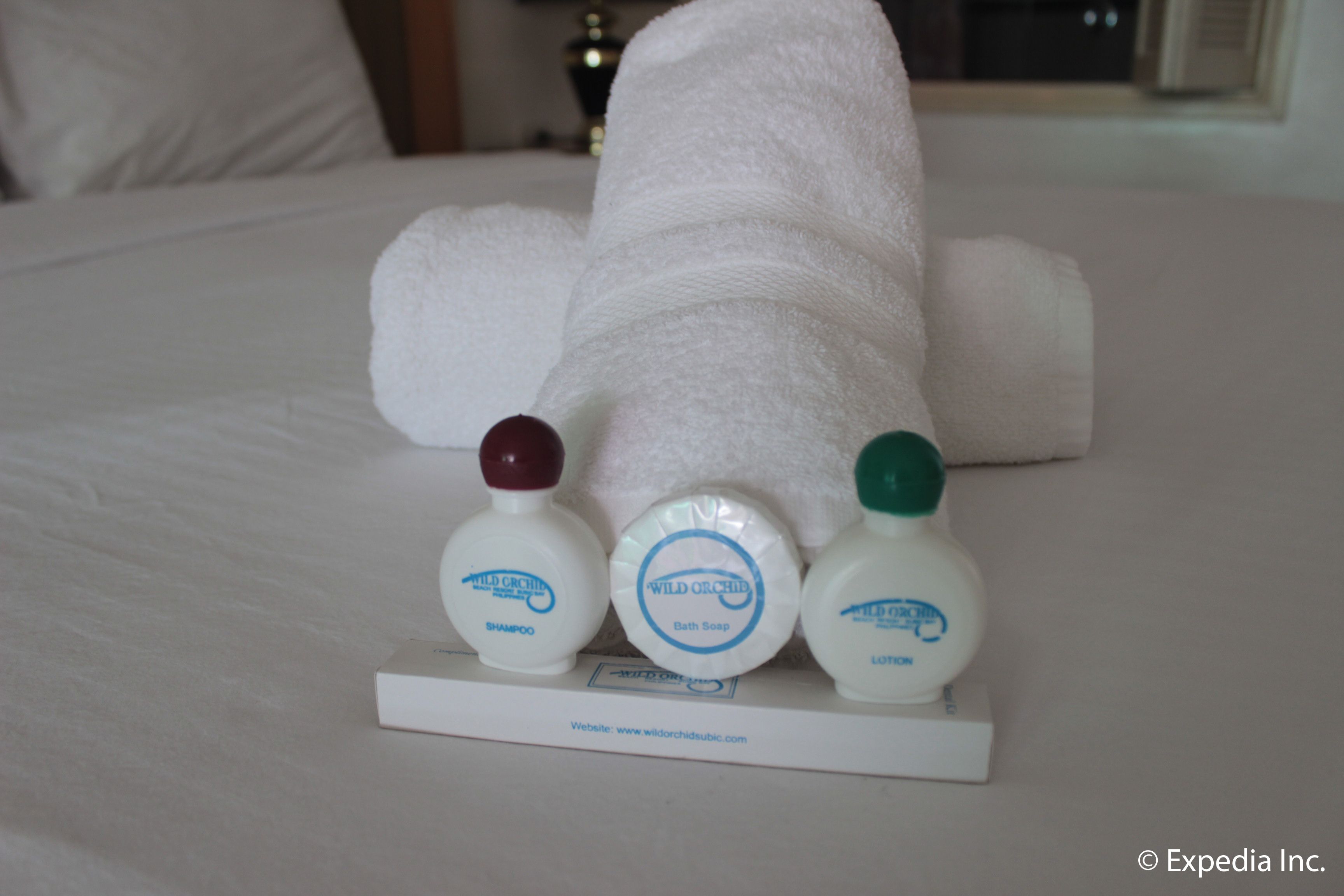 A neatly made bed with a folded white towel and three bottles of Wild Orchid products: shampoo, bath soap, and lotion.