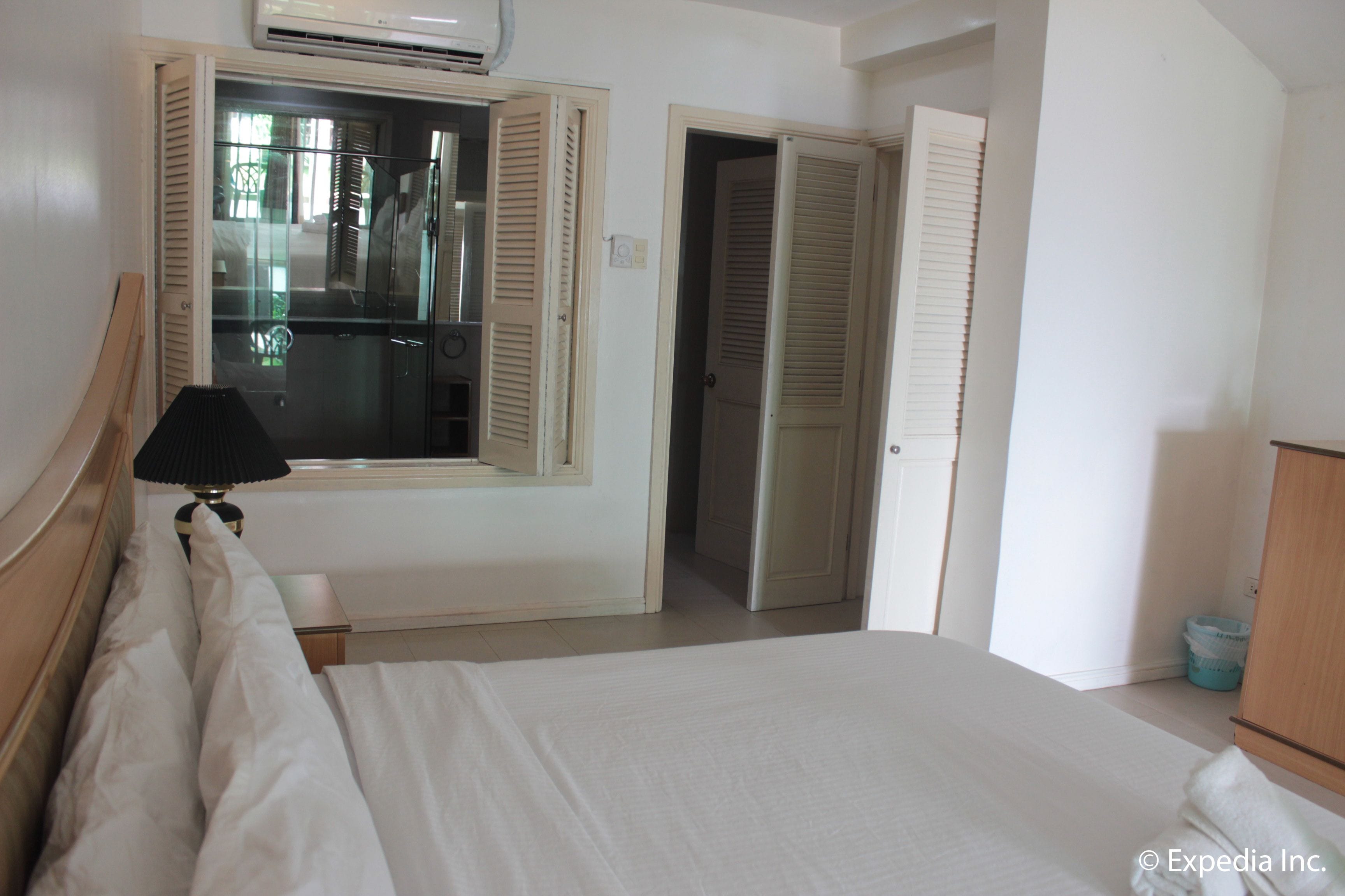 A hotel room with a bed, a bedside table with a lamp, a window with shutters, and a door leading to another room.