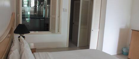 A hotel room with a bed, a bedside table with a lamp, a window with shutters, and a door leading to another room.