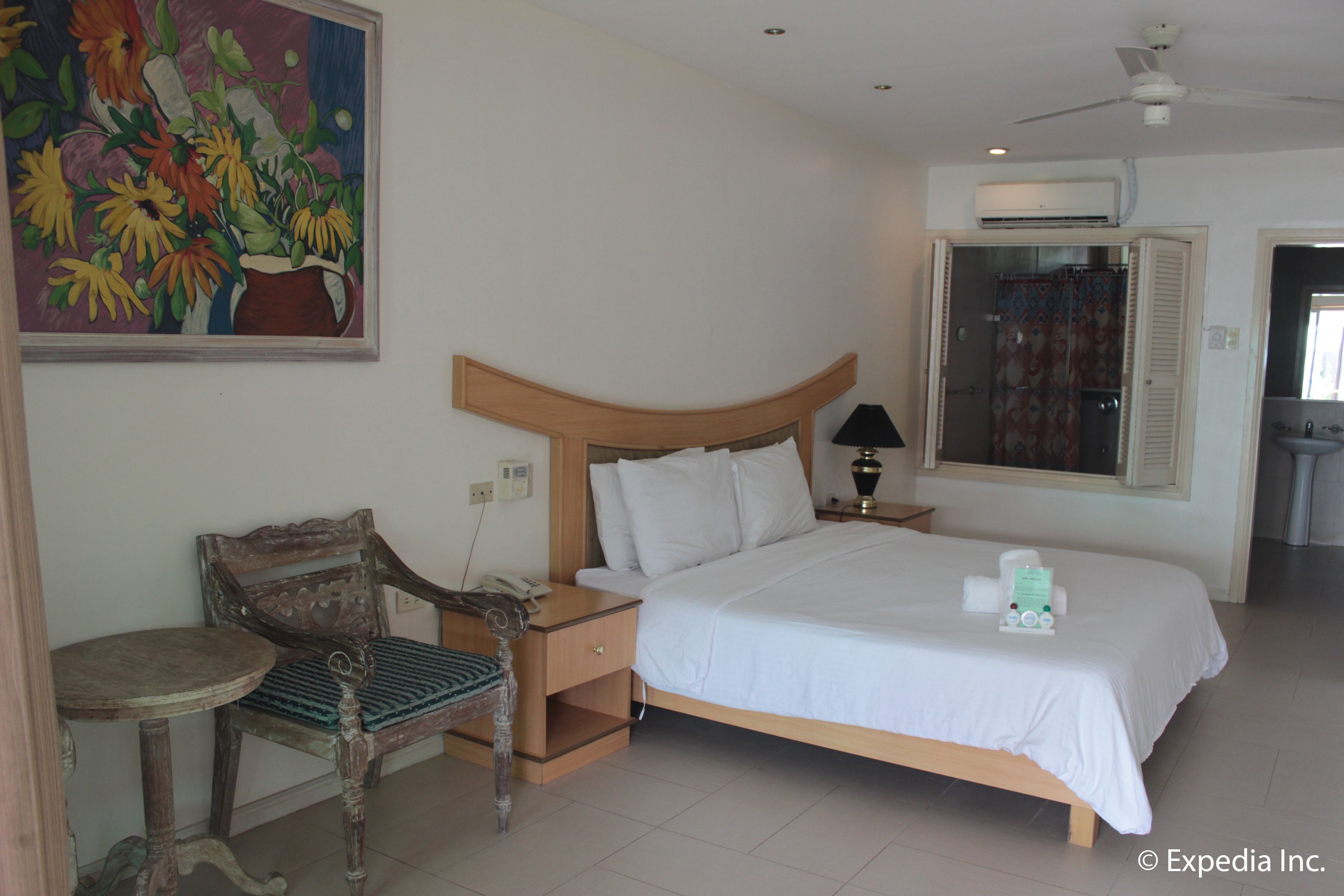 Une chambre d’hôtel avec un lit, une table de chevet en bois, une chaise, une petite table, un tableau, un appareil de climatisation et une salle de bain visible à travers une porte ouverte.