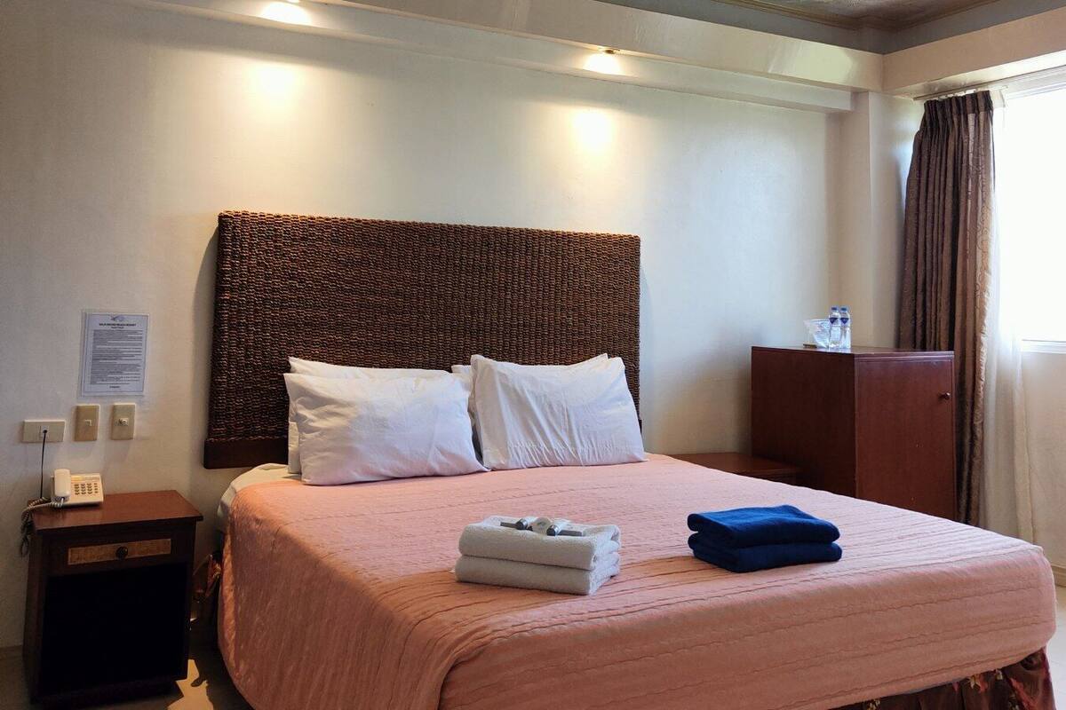 A hotel room with a large bed, a wooden headboard, a nightstand with a phone, a desk with a lamp, and a window with curtains.