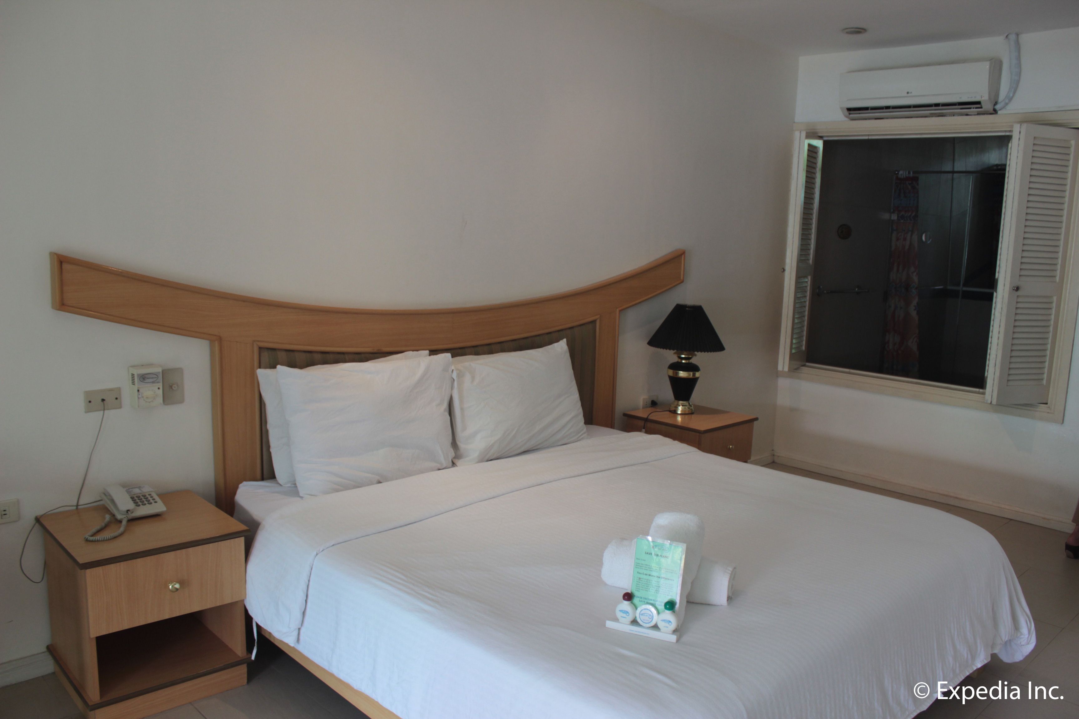 Habitación de hotel con cabecera de madera, lámpara de noche, teléfono en la mesita y una cama bien hecha con artículos de aseo.