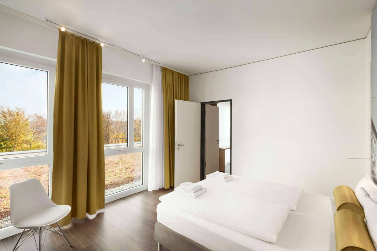 Quarto de hotel moderno com uma cama grande, uma cadeira e vista para árvores pela janela.