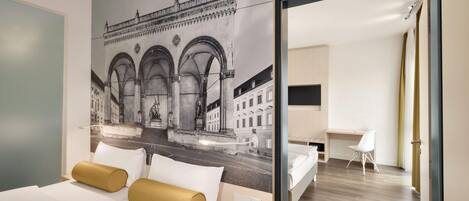 Habitación de hotel con cama, escritorio y vista a la calle a través de un ventanal grande.