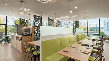 Ein modernes Restaurant mit hohen Tischen, gepolsterten grünen Sitzbänken, großen Fenstern und einem geometrischen Deckenmuster.