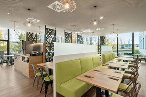 A modern restaurant with high tables and green cushioned booths, large windows, and a geometric ceiling design.