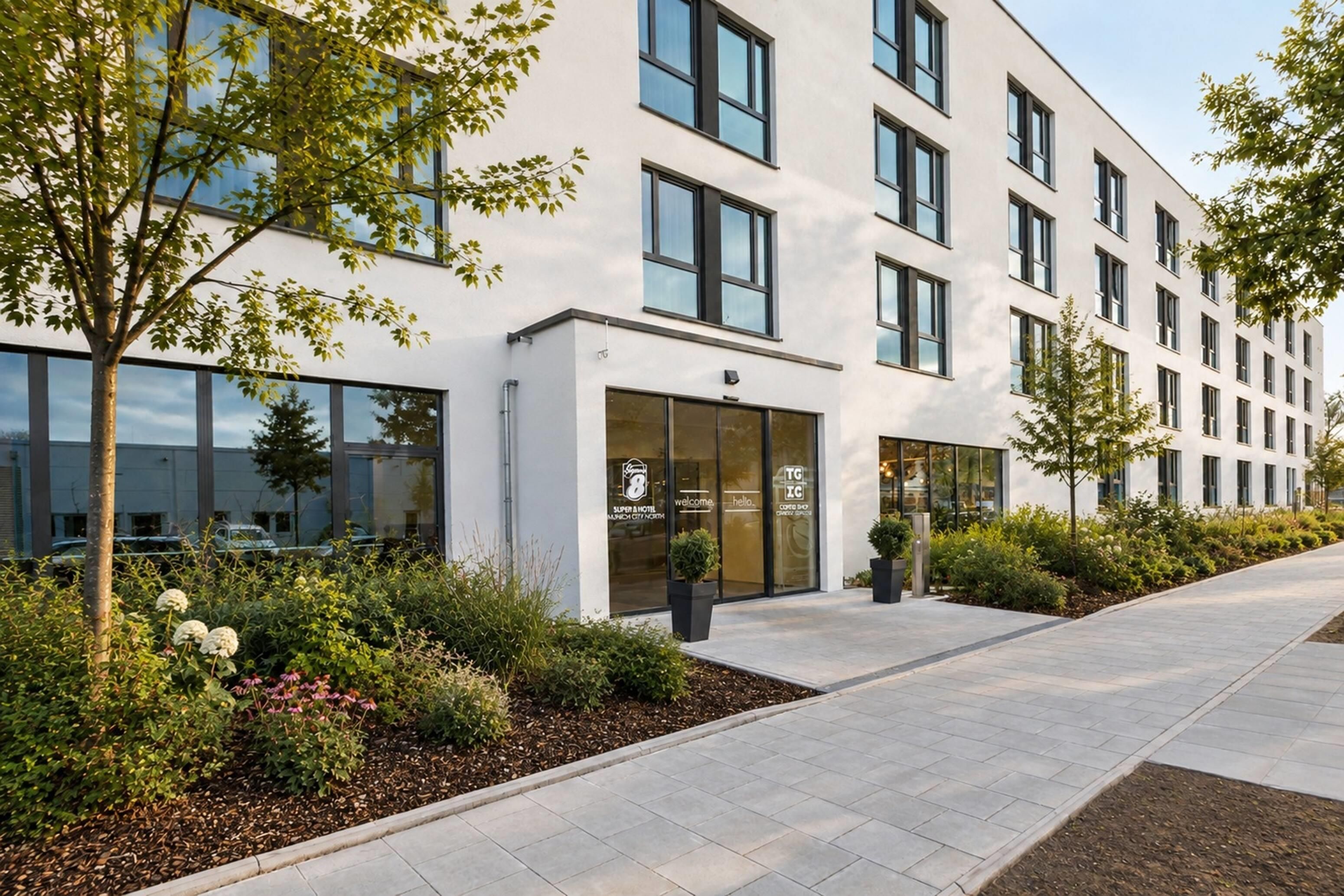 Un hôtel moderne à la façade blanche, doté de grandes fenêtres et d’une entrée agrémentée d’un aménagement paysager.