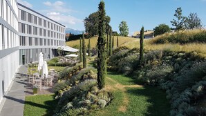 Un edificio moderno con un patio que incluye mobiliario exterior y un jardín ajardinado con arbustos y árboles diversos.
