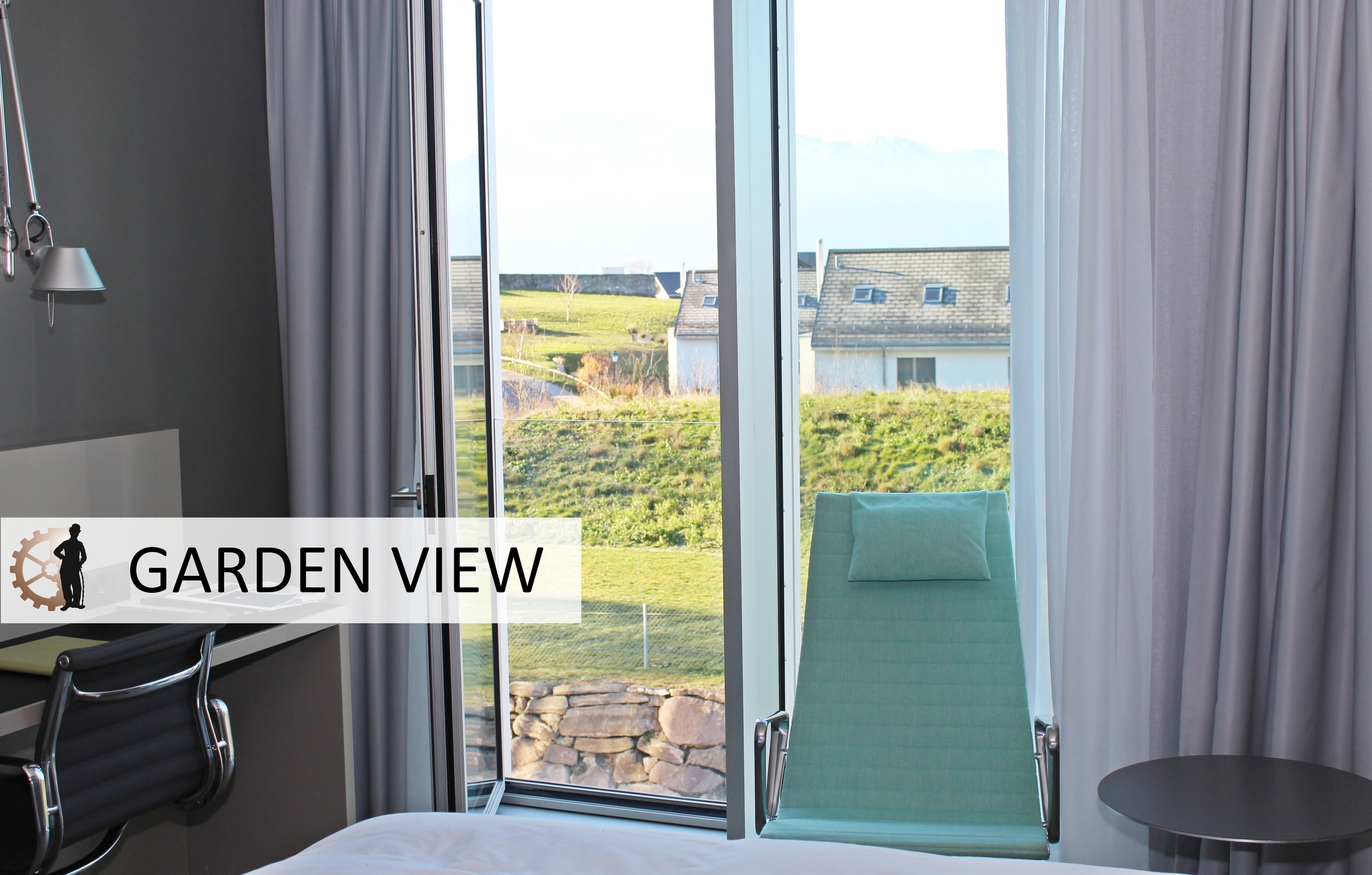 Ein Hotelzimmer mit großem Fenster und Blick auf einen Garten, inklusive Bett, Stuhl und kleinem Tisch.