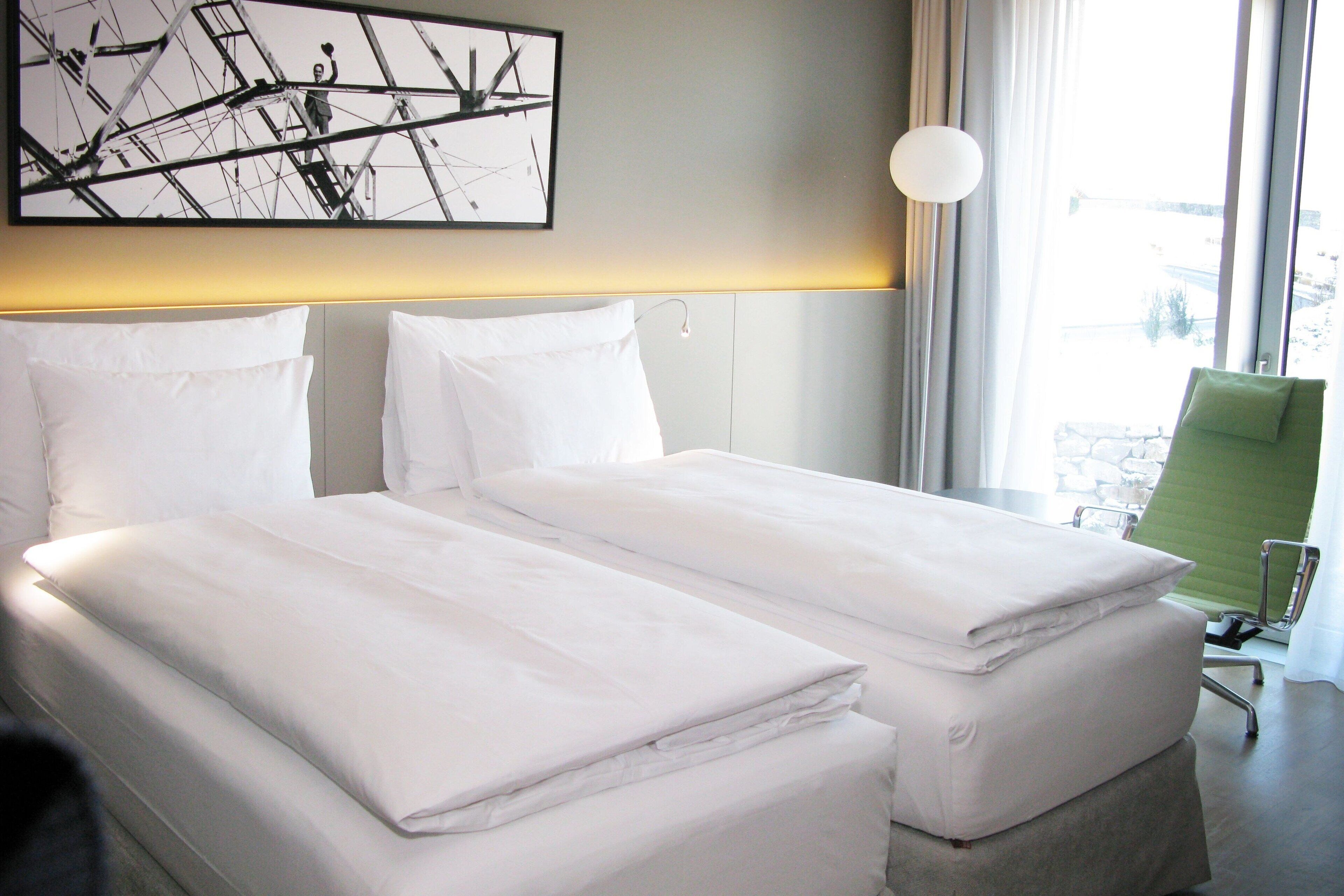 A hotel room with two beds, a green chair, and a large window with curtains.