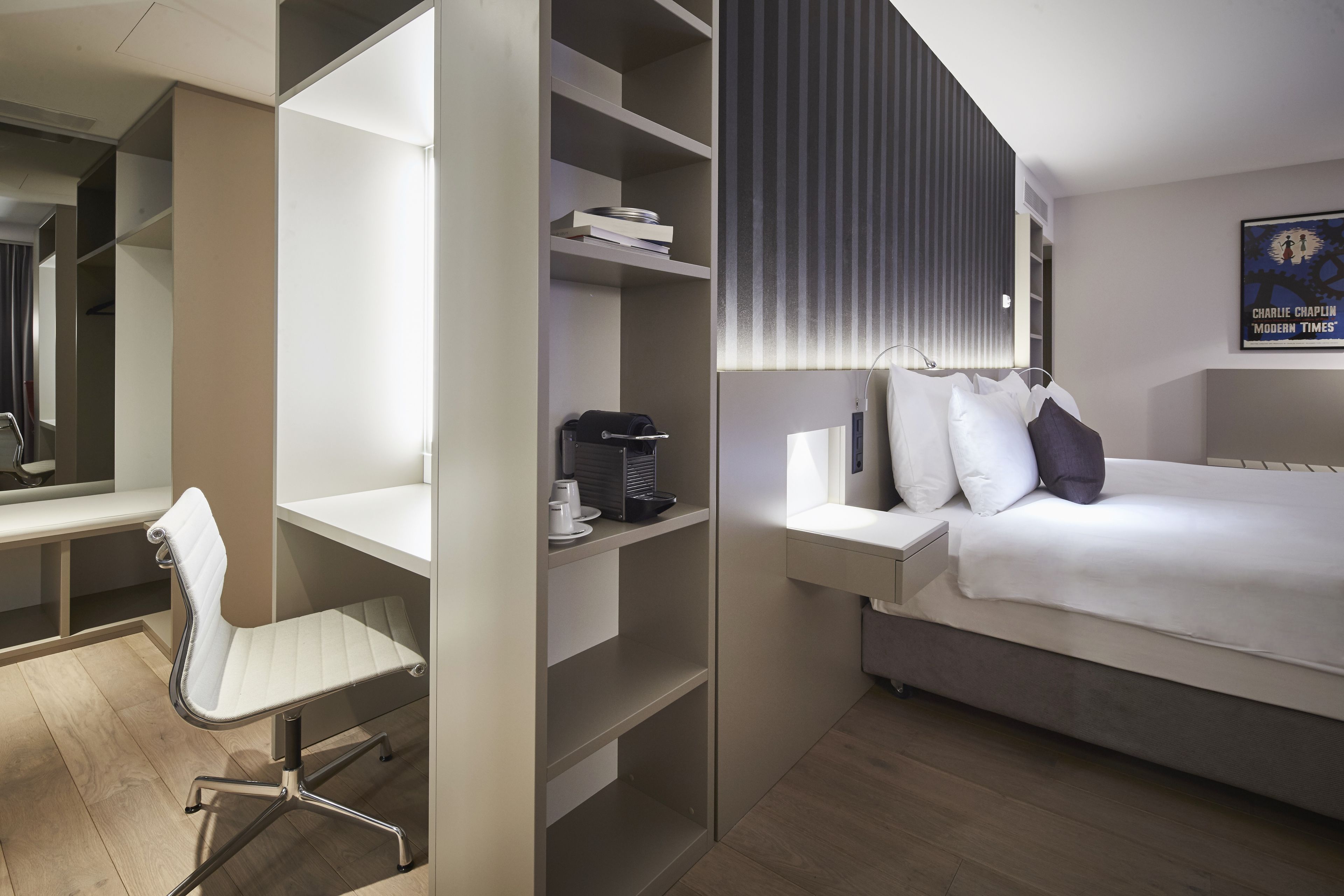 A modern hotel room with a bed, a desk, a chair, and built-in shelving.