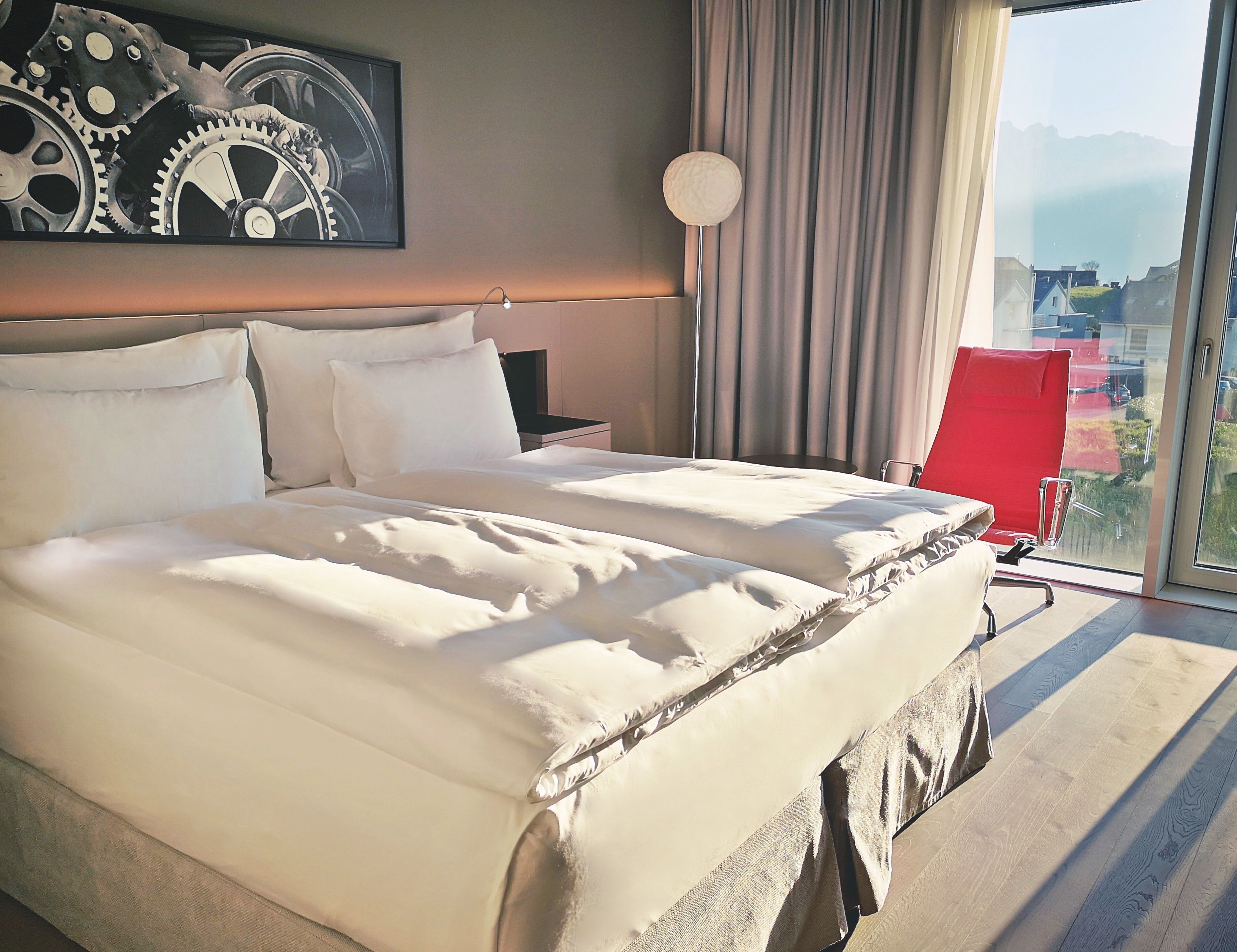 A hotel room with a large bed, a nightstand, a lamp, and a view of the city through the window.