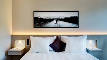 Ein Hotelzimmer mit einem Bett, zwei Nachttischen und einem gerahmten Foto von einem Paar, das auf einer Straße mit Bergen im Hintergrund spazieren geht.