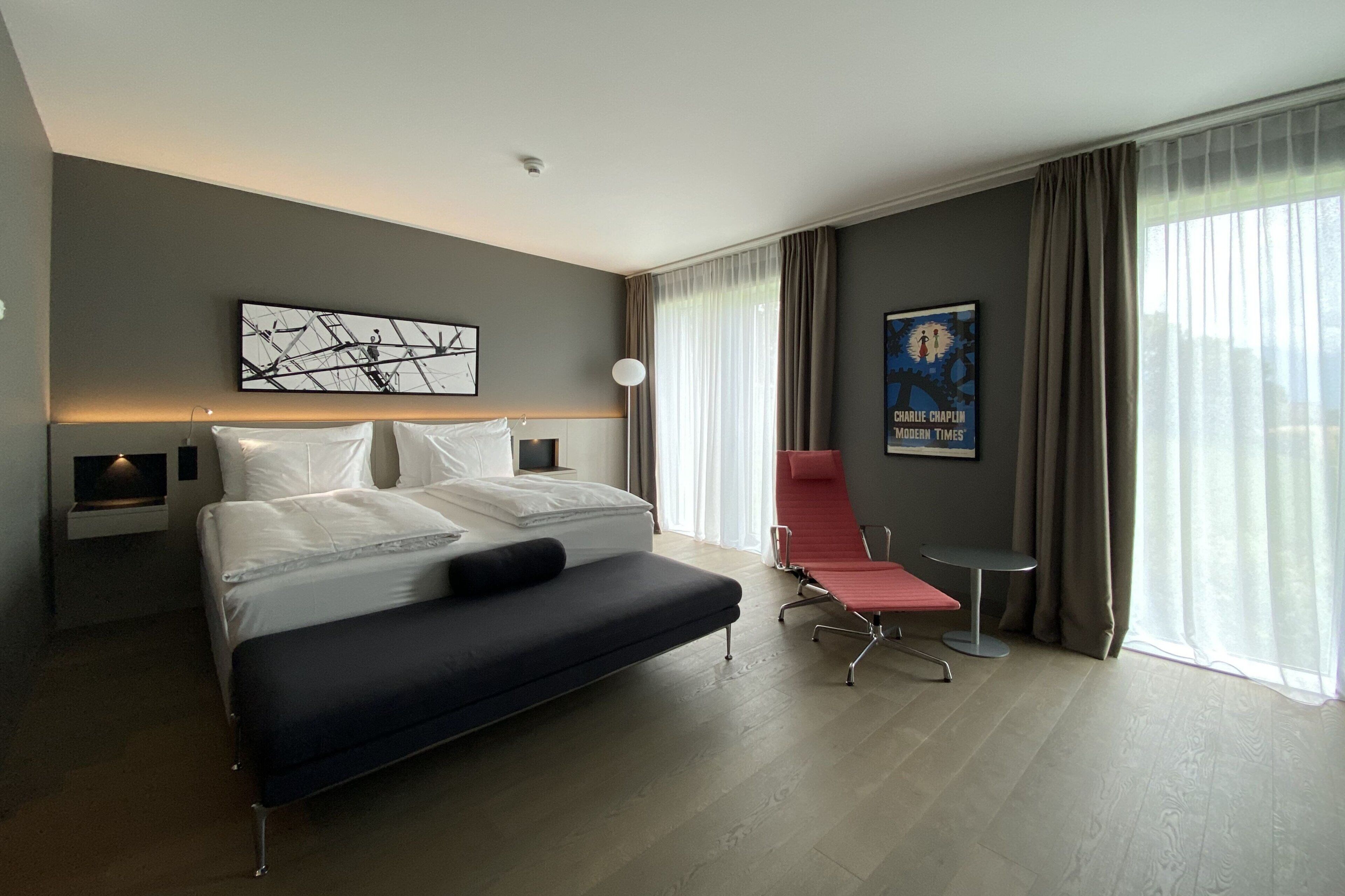 A modern hotel room with a large bed, a bedside table, a reading chair, and a poster on the wall.