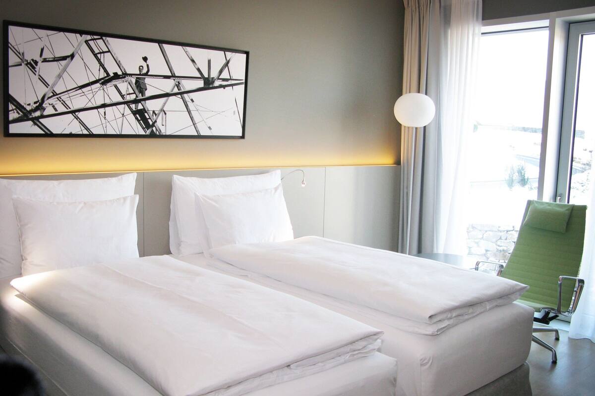 Une chambre d’hôtel avec deux lits, un fauteuil vert, une grande fenêtre et un tableau au mur.