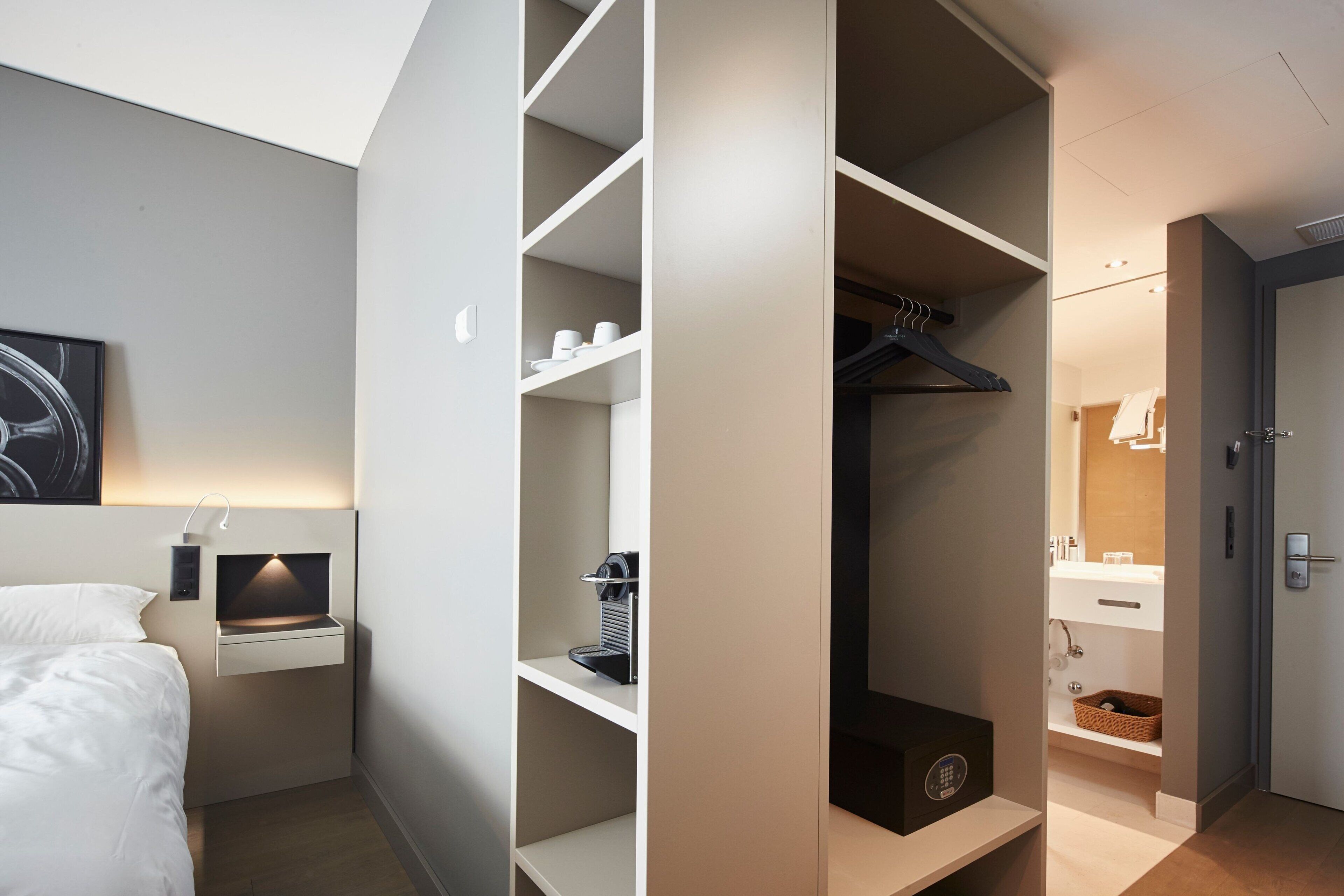 Una habitación de hotel moderna con un armario ordenado, una mesita de noche con lámpara y una cama grande.