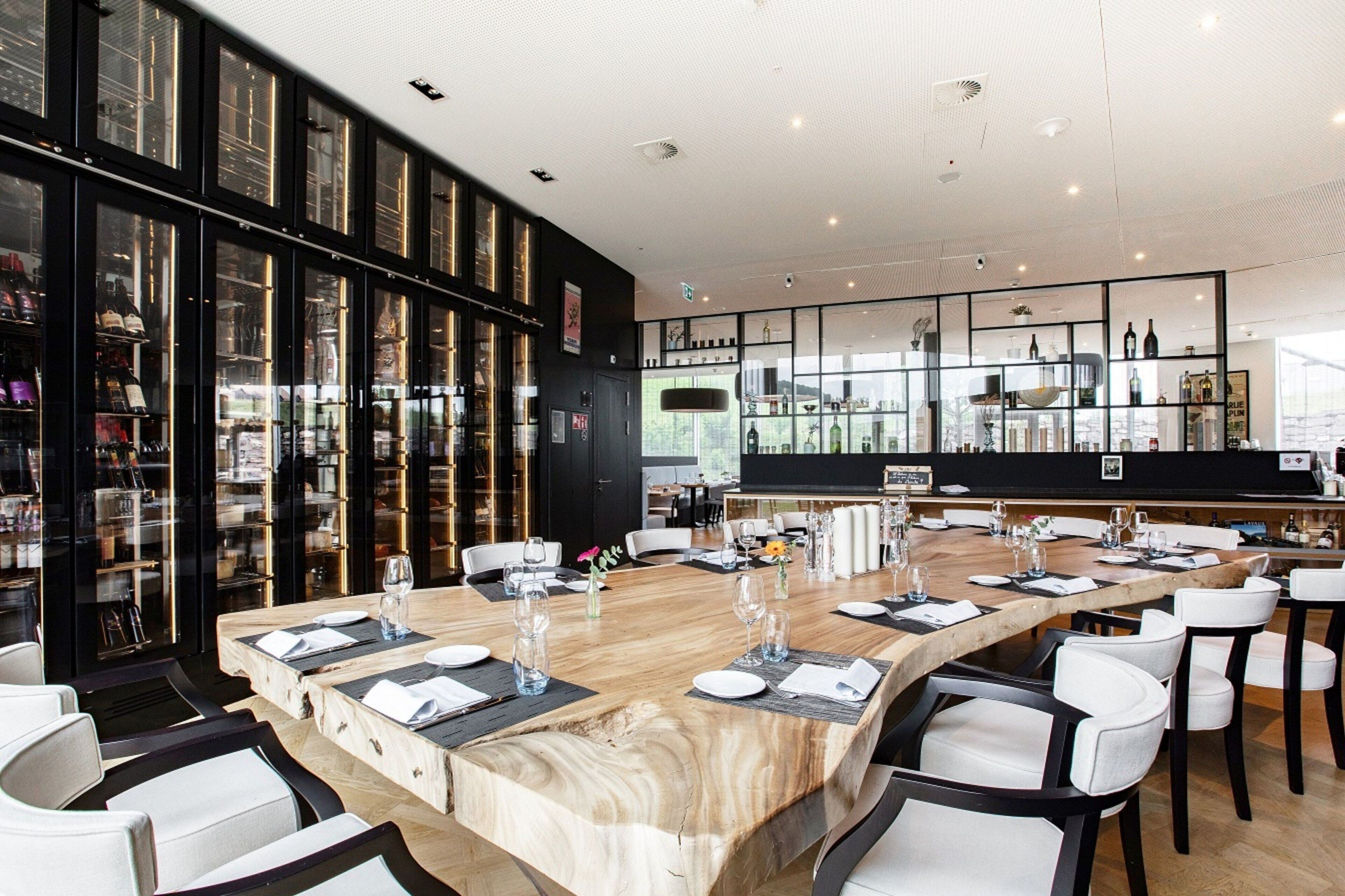 A modern restaurant with a long wooden table set for dining, surrounded by white chairs.