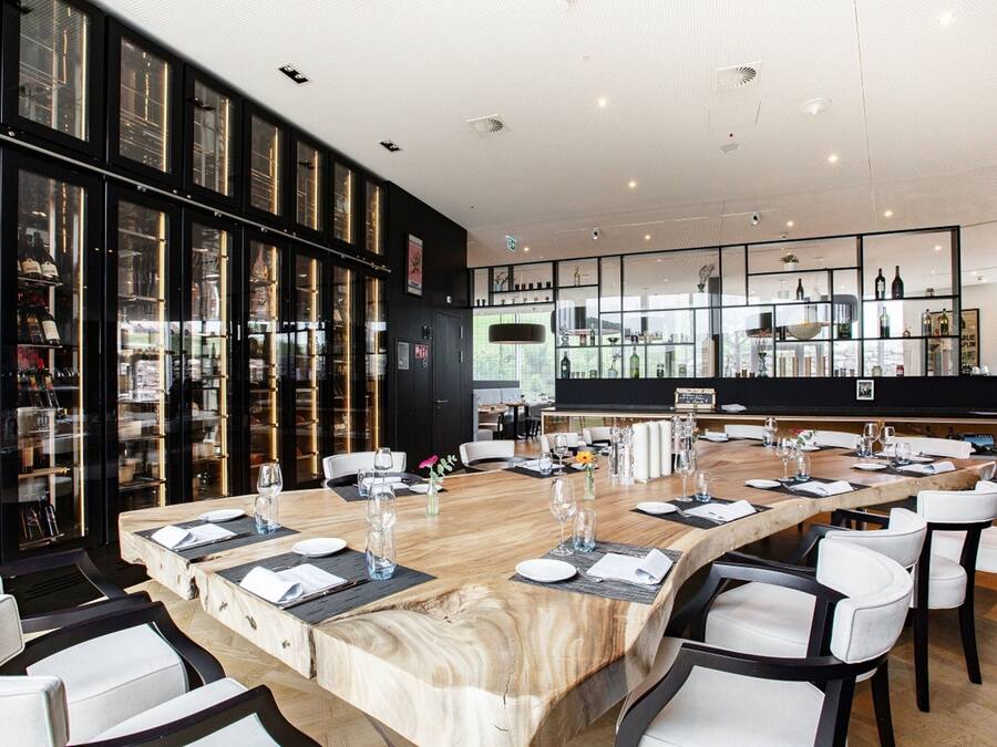 A modern restaurant with a long wooden table set for dining, surrounded by white chairs.