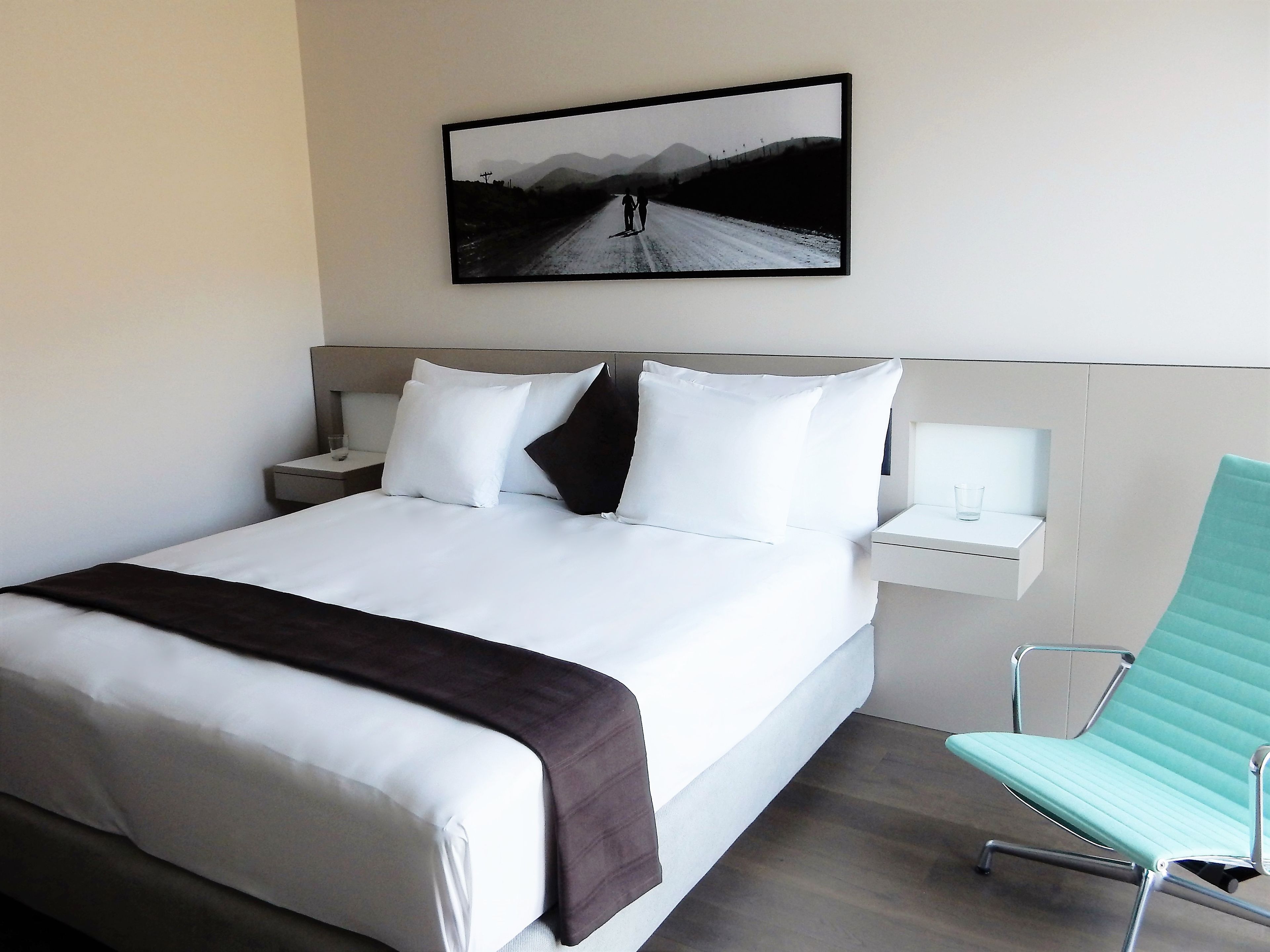 Une chambre d’hôtel avec un grand lit, une table de chevet, une petite table de nuit avec une lampe, une chaise et un tableau encadré au mur.