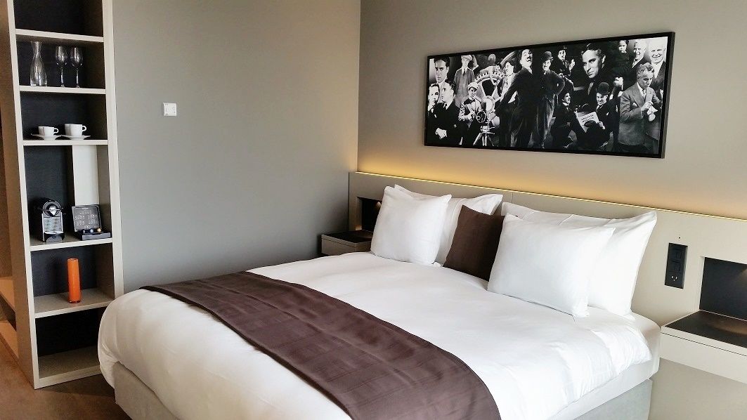 Ein ordentlich eingerichtetes Hotelzimmer mit einem großen Bett, einem Nachttisch, einem Regal mit Gläsern und einem Radio sowie einem an der Wand hängenden Bild.