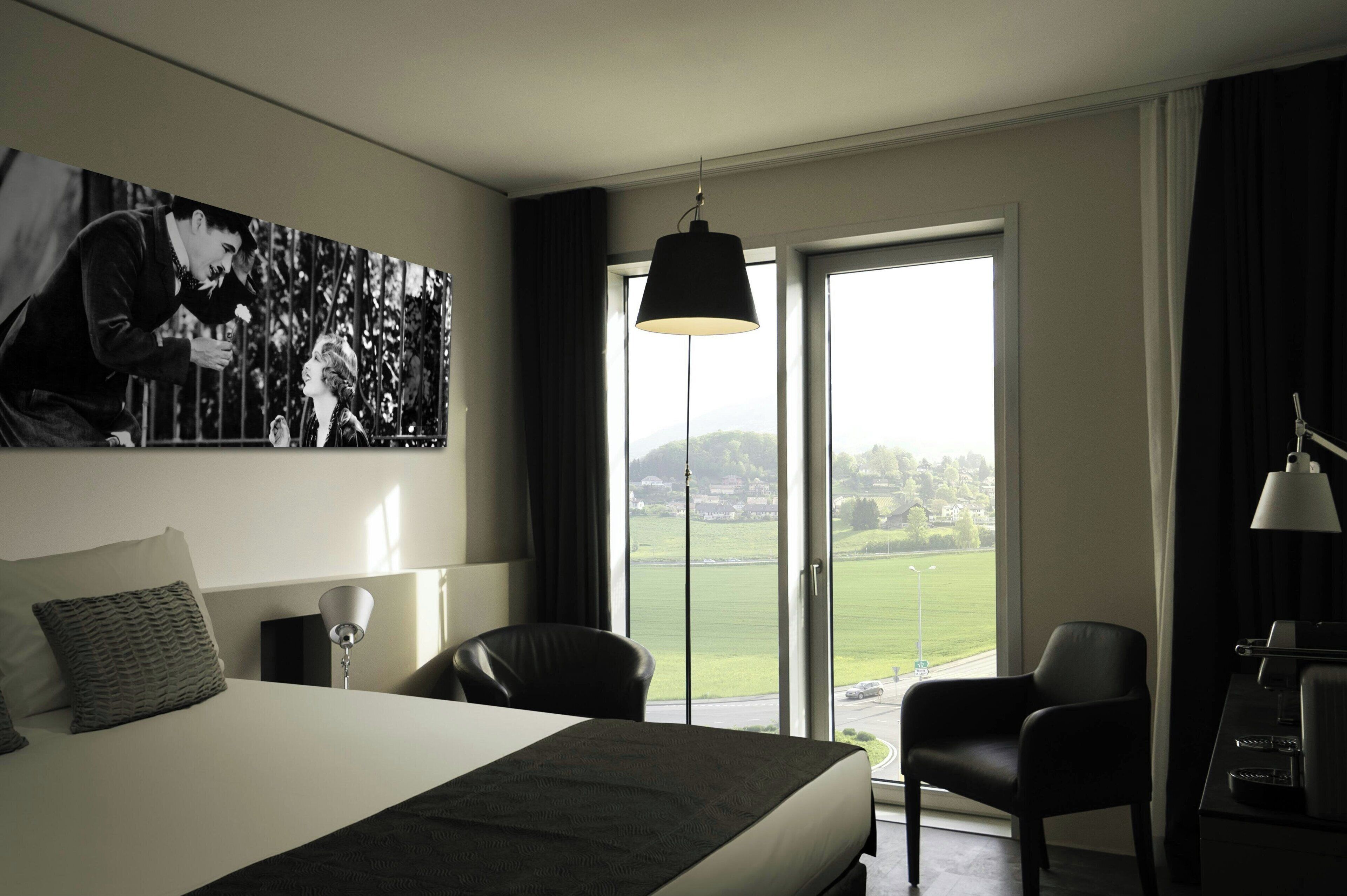 A hotel room with a large bed, a desk with a lamp, a chair, and a view of a green landscape through the window.