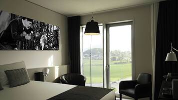 A hotel room with a large bed, a desk with a lamp, a chair, and a view of a green landscape through the window.