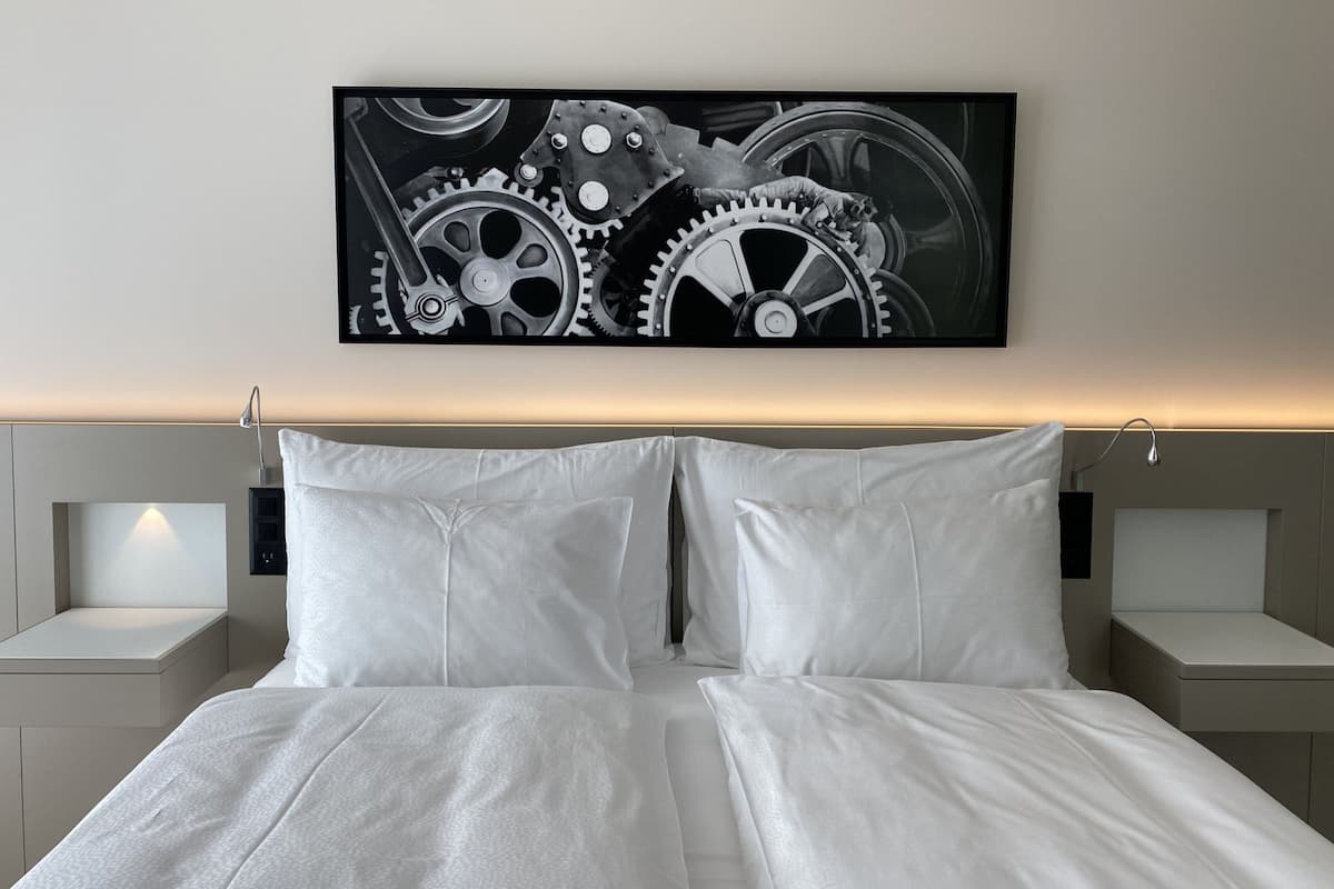 Une chambre moderne avec un grand lit, une literie blanche et une œuvre d’art représentant des engrenages, accrochée au mur.