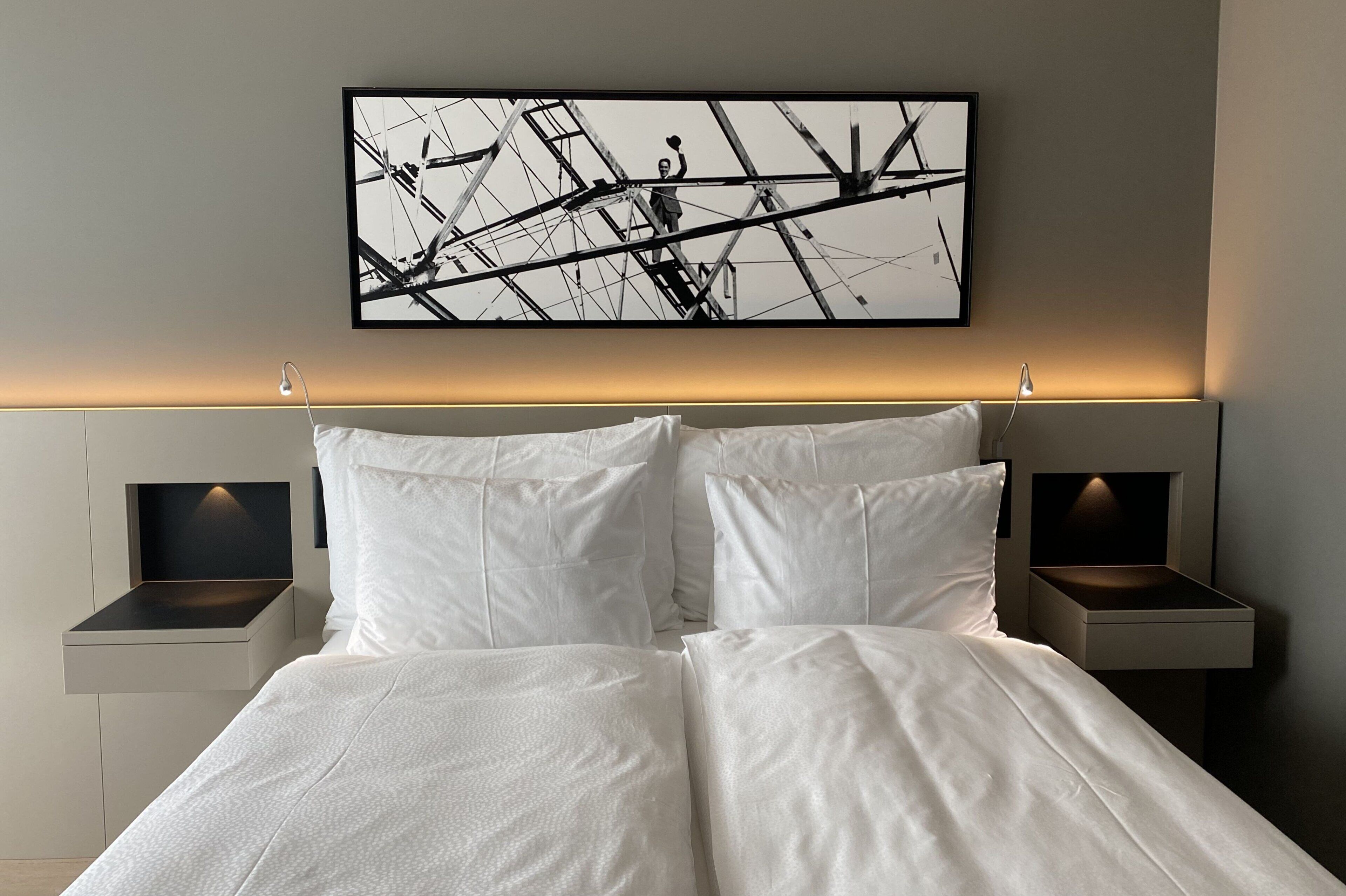 A modern hotel room with a large bed, white linens, two bedside tables with lamps, and a framed abstract artwork on the wall.