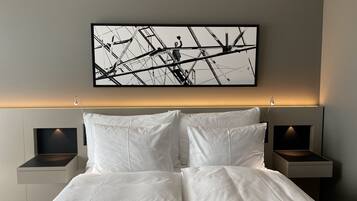 A modern hotel room with a large bed, white linens, two bedside tables with lamps, and a framed abstract artwork on the wall.